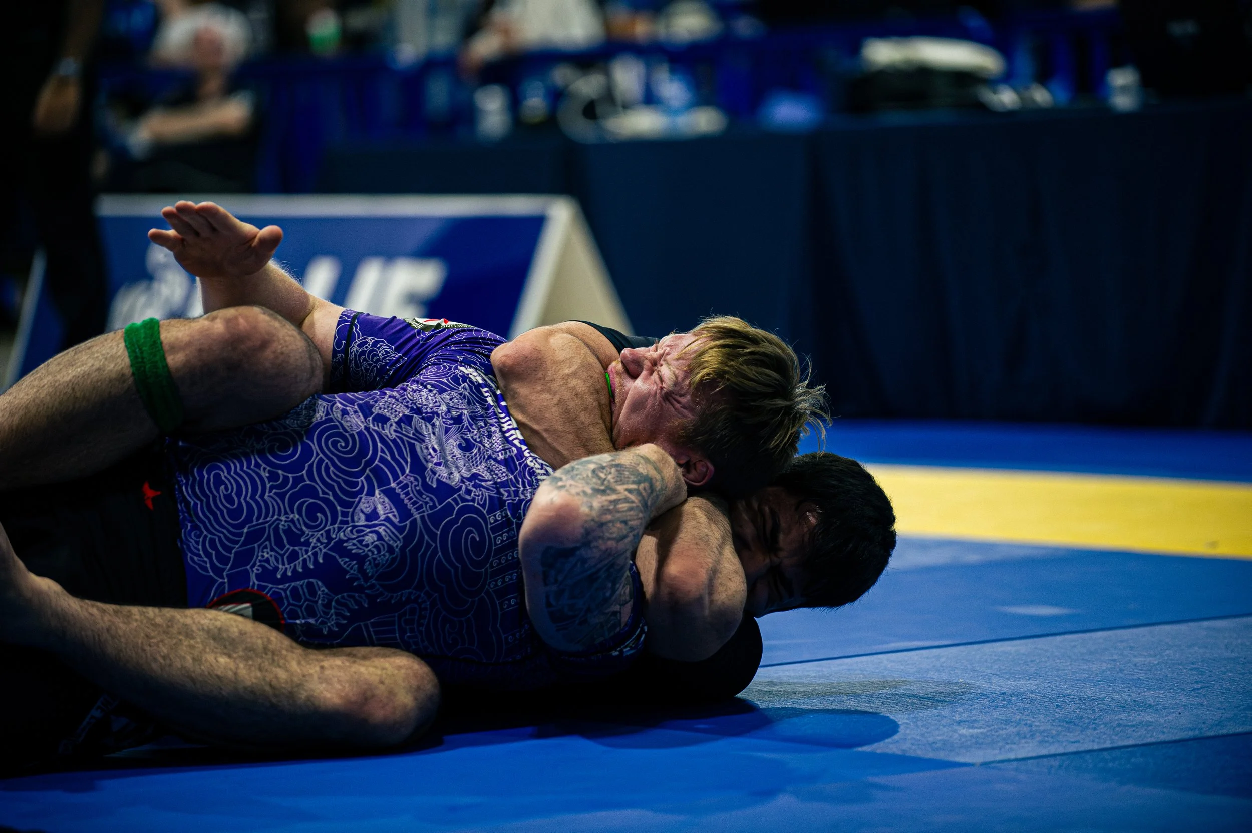 Two male wrestlers engaged in a grappling match on a blue mat at a wrestling competition, with one wrestler applying a hold on the other.