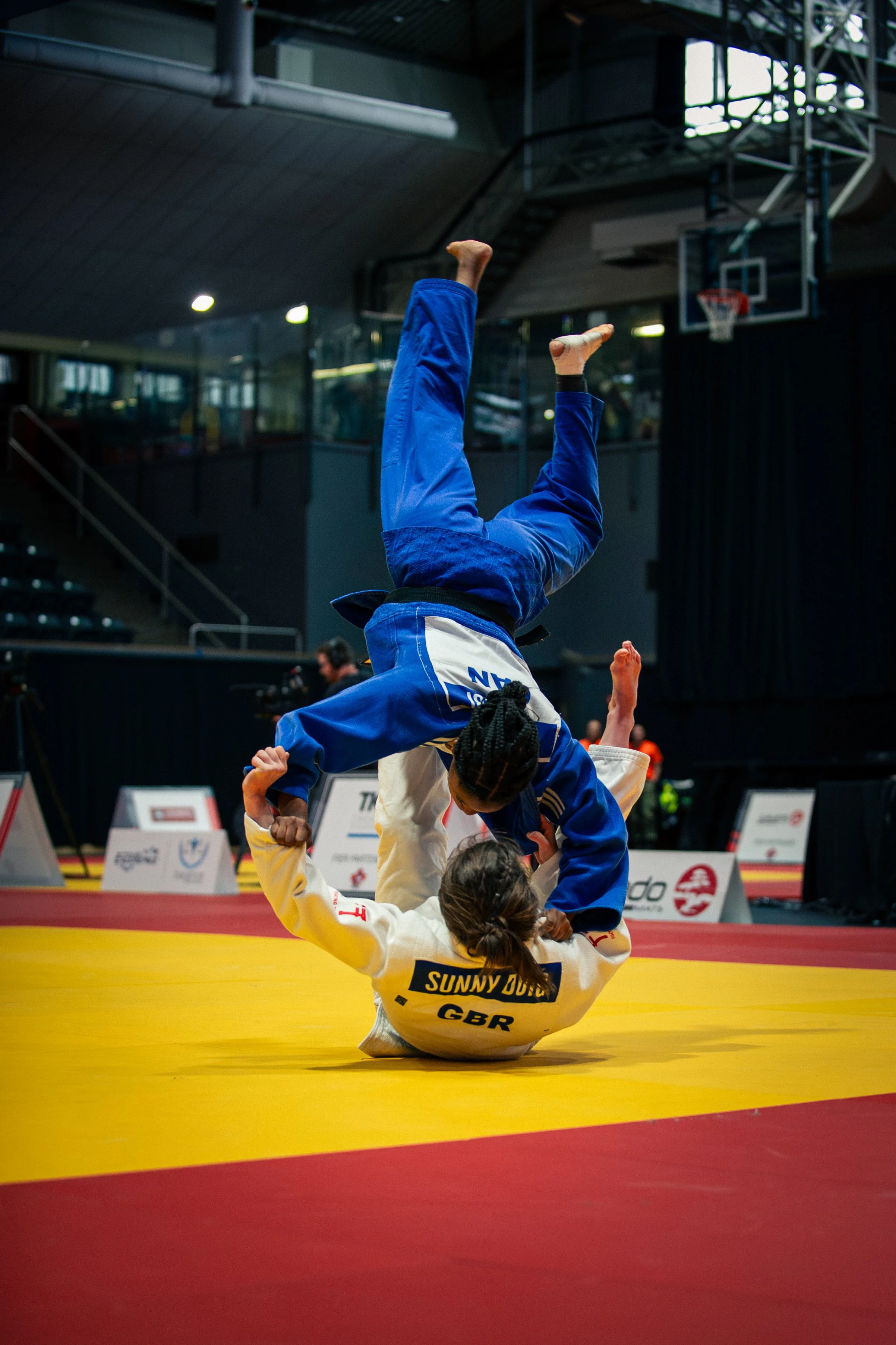 A martial arts competitor wearing a yellow gi with black text and a blue gi with white text is performing a throw during a competition on a yellow mat. The competitor's gi has the name "SUNNY JONES" and "GBR" printed on the back. Two other competitor