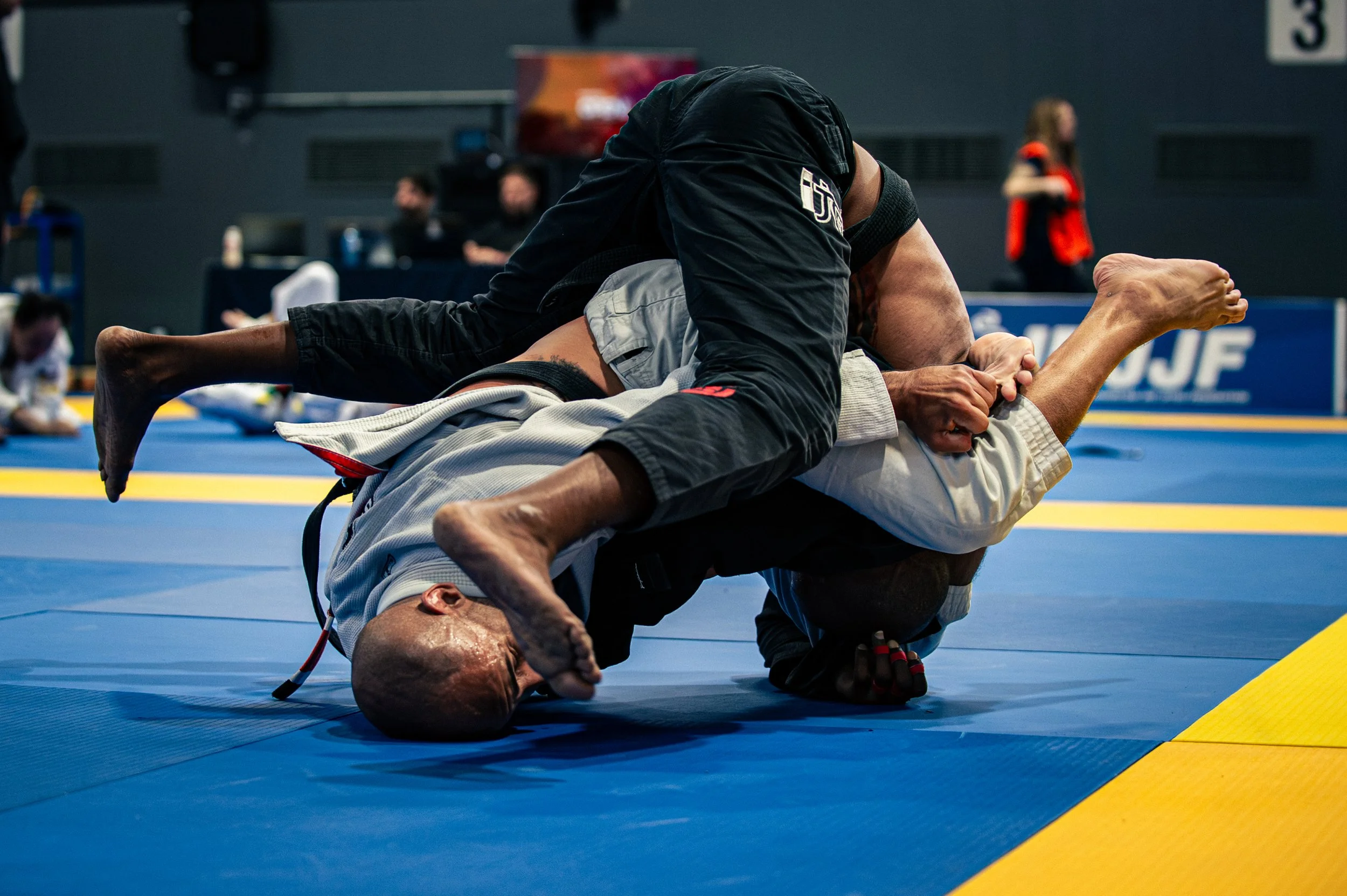 Two Brazilian Jiu-Jitsu practitioners engaged in a grappling match on a blue mat, with one person in a black gi and the other in a white gi, in an indoor sports facility. IBJJF Montreal Jiu-JItsu