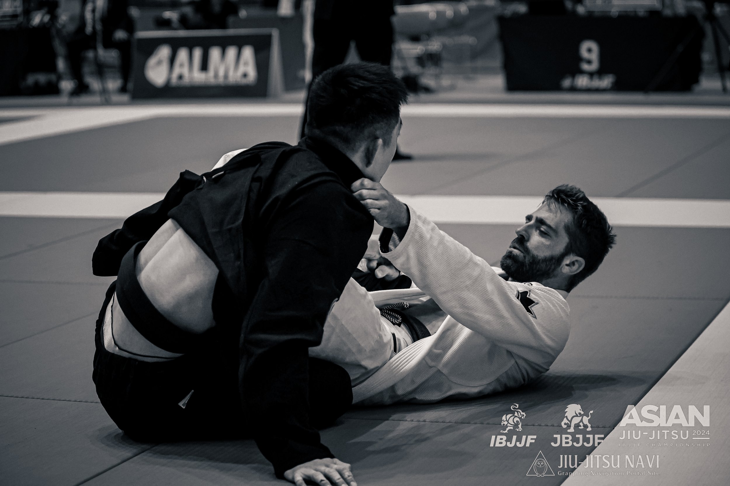 Two men practicing Brazilian Jiu-Jitsu on the mat, with one man on top and the other lying on the ground, at Chiba Port Arena, Japan , with banners and logos in the background.