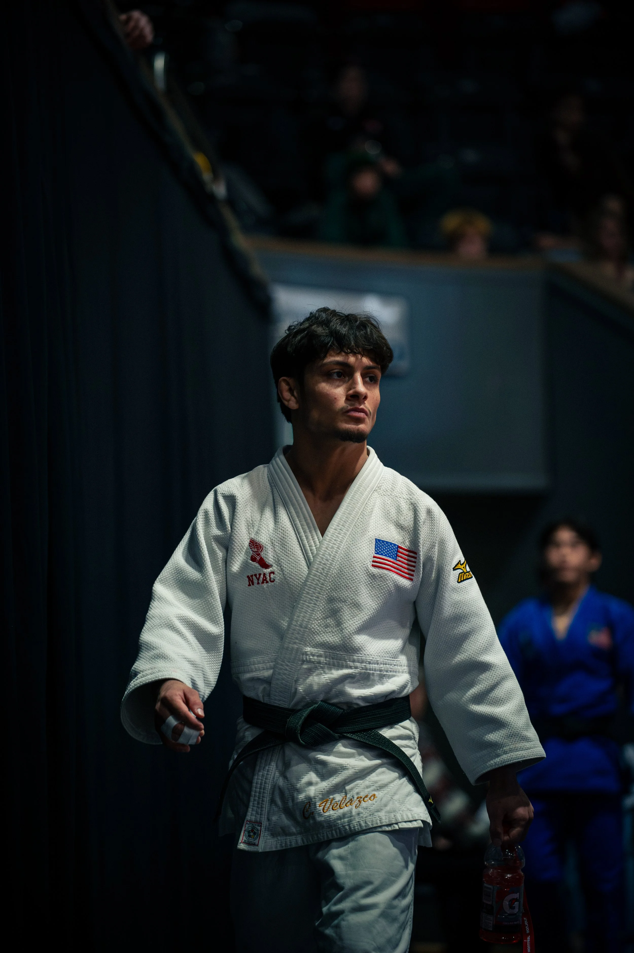 A male martial artist in a white gi with patches and a black belt, holding a water bottle, standing in a dimly lit room with other competitors in the background.