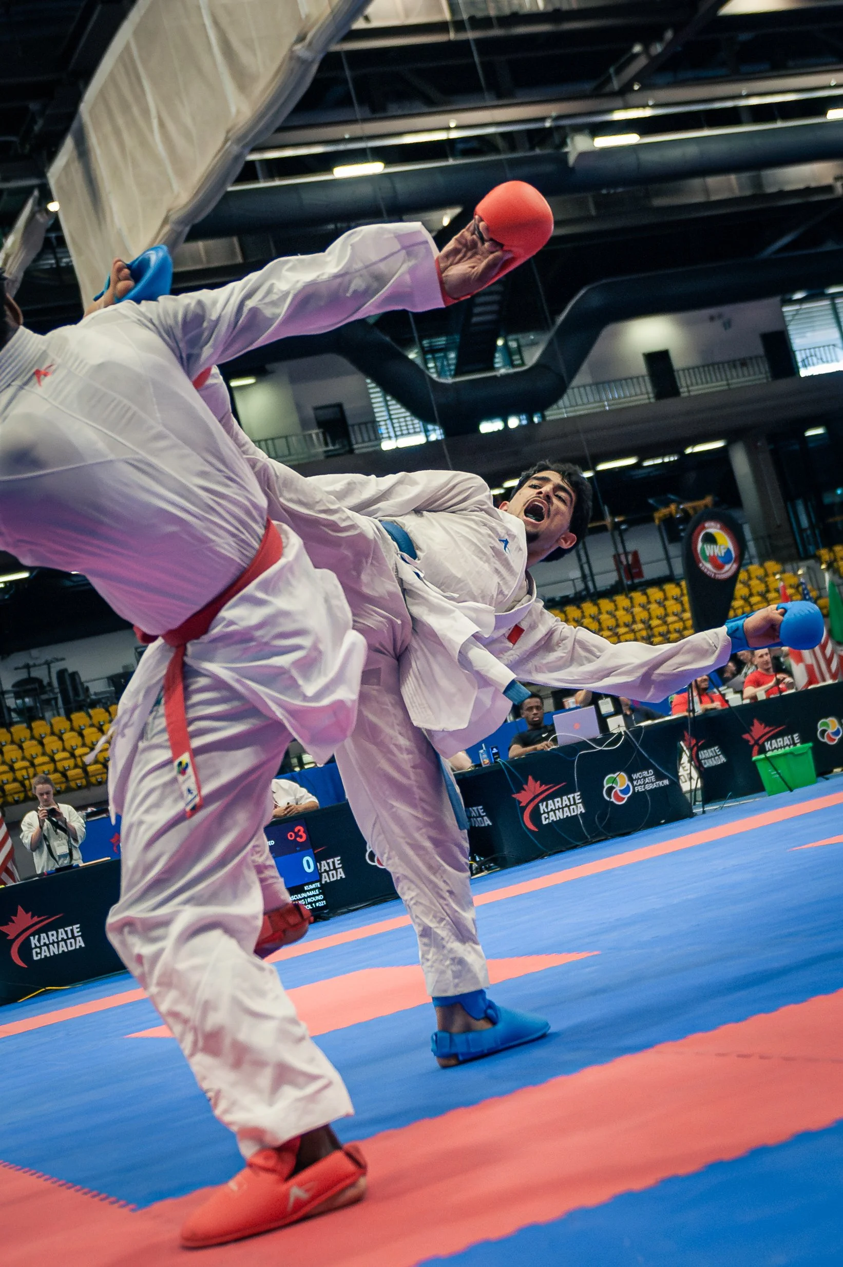 Two martial artists competing in a karate match on a blue and orange mat in a large indoor arena, with spectators and event banners visible in the background. montreal international open
