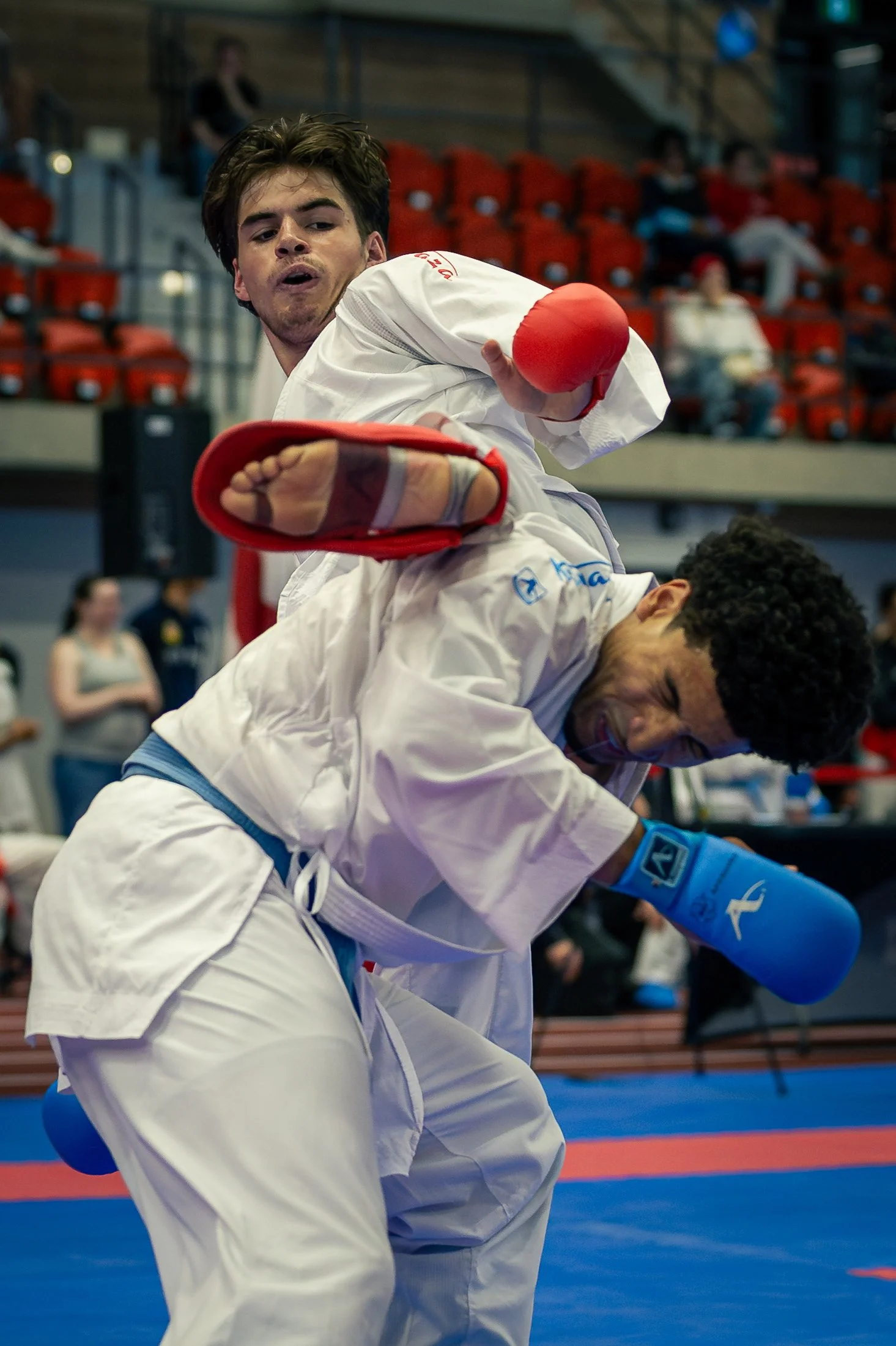 Two martial artists in white uniforms practicing karate in a gymnasium, with red and blue sparring mats and spectators in the background.