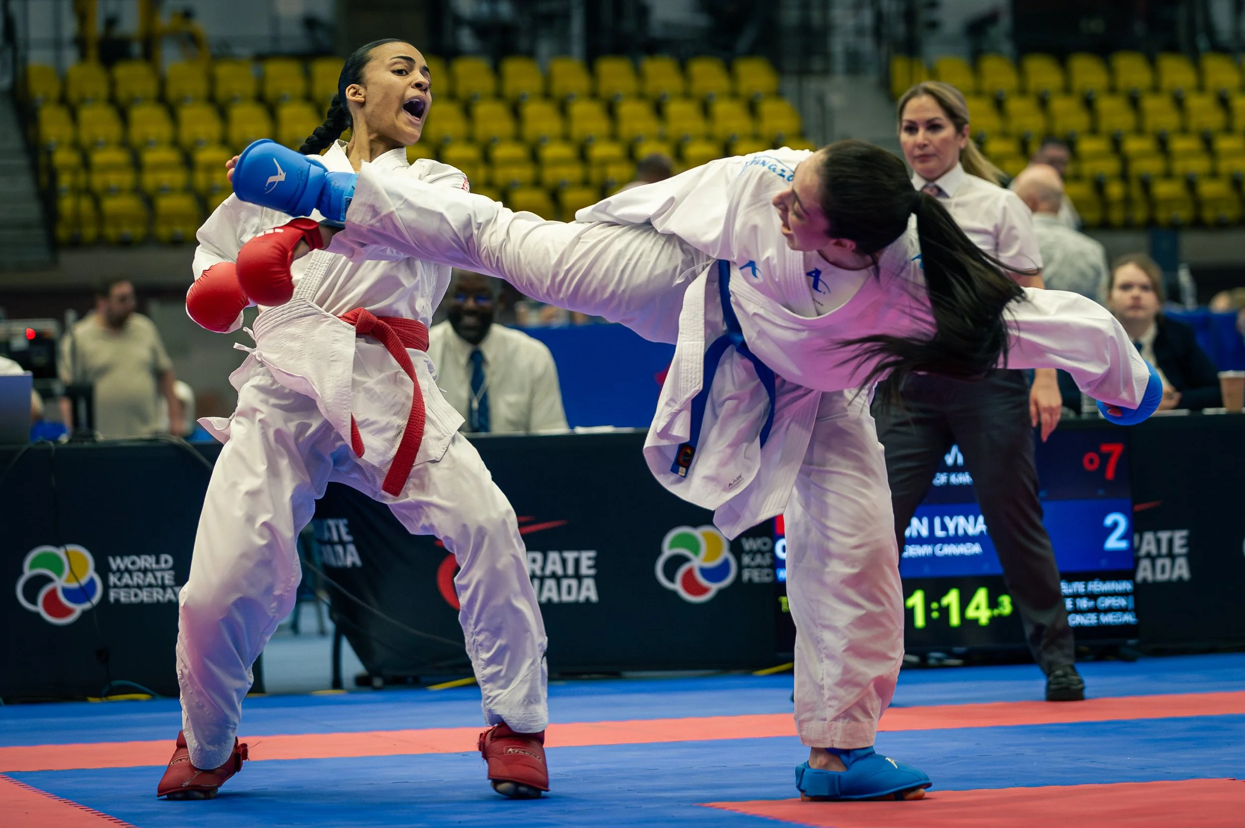 Two female karate competitors in white gis with colored belts engaging in a match on a blue and red mat, with one executing a kick towards the other's face, at a tournament with yellow seating and digital scoreboard.