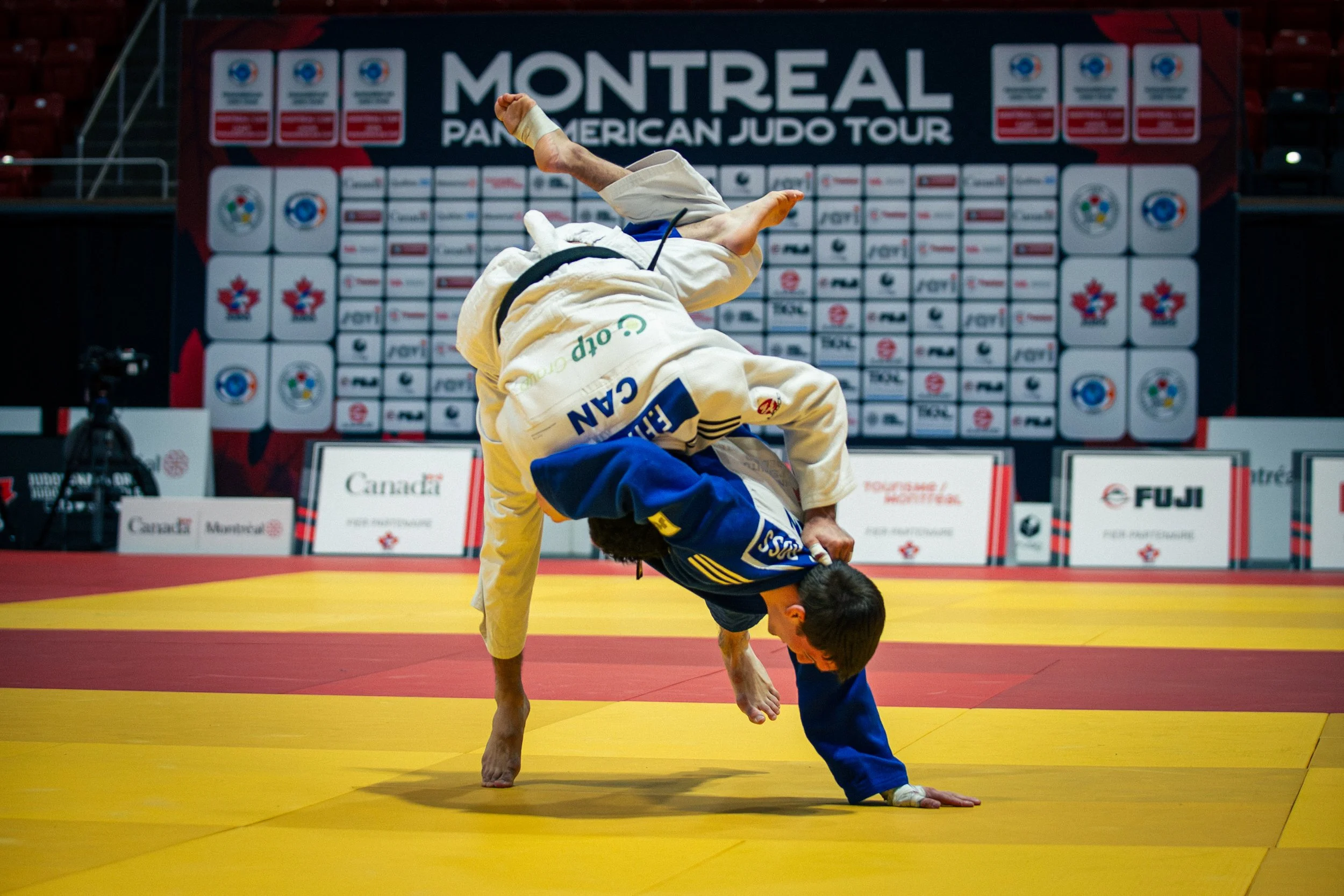 Two judo athletes competing on a yellow mat during a match at the Montreal Pan-American Judo Tour, with a backdrop of sponsor logos and event signage.