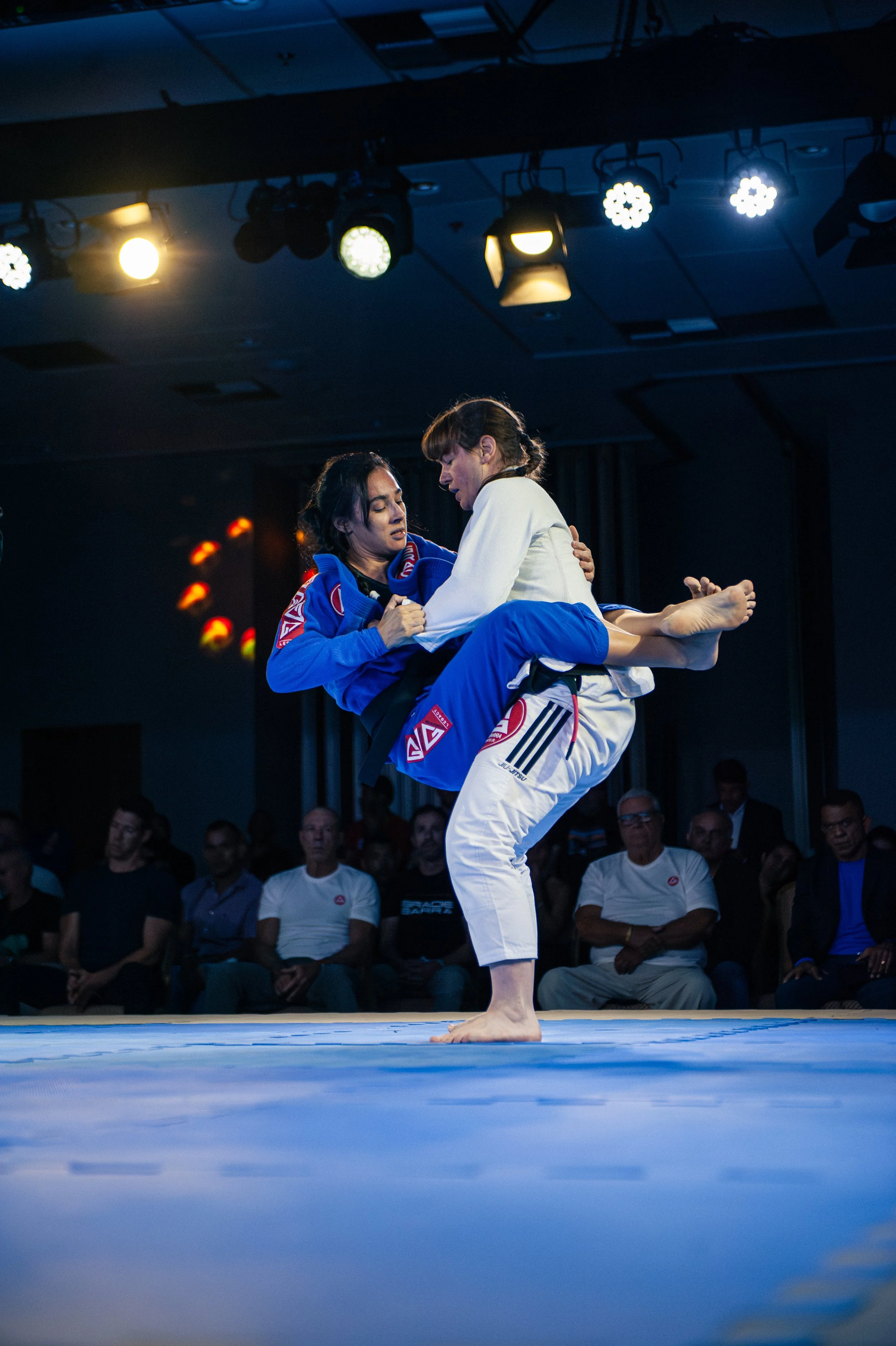 Two women are practicing Brazilian Jiu-Jitsu on a blue mat, with one woman executing a throw on the other. The instructor and an audience of spectators watch in the background, with bright overhead lights illuminating the scene. Carlos Gracir JR, Rio