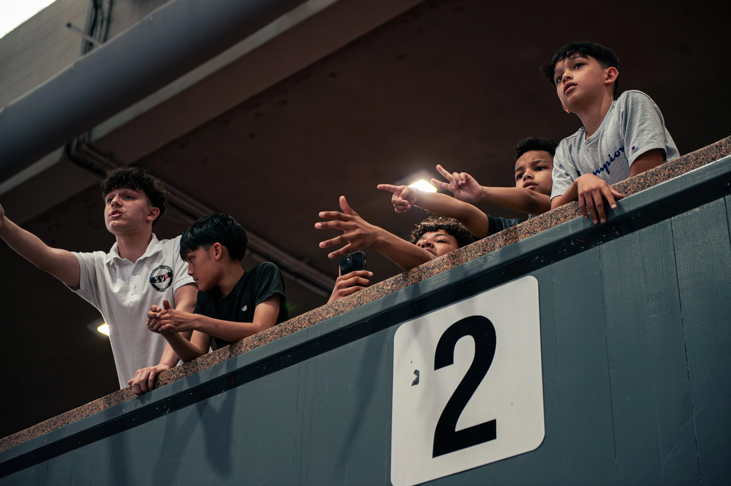 Group of young boys leaning over a balcony in an indoor stadium or arena, engaged with something below or in front of them.