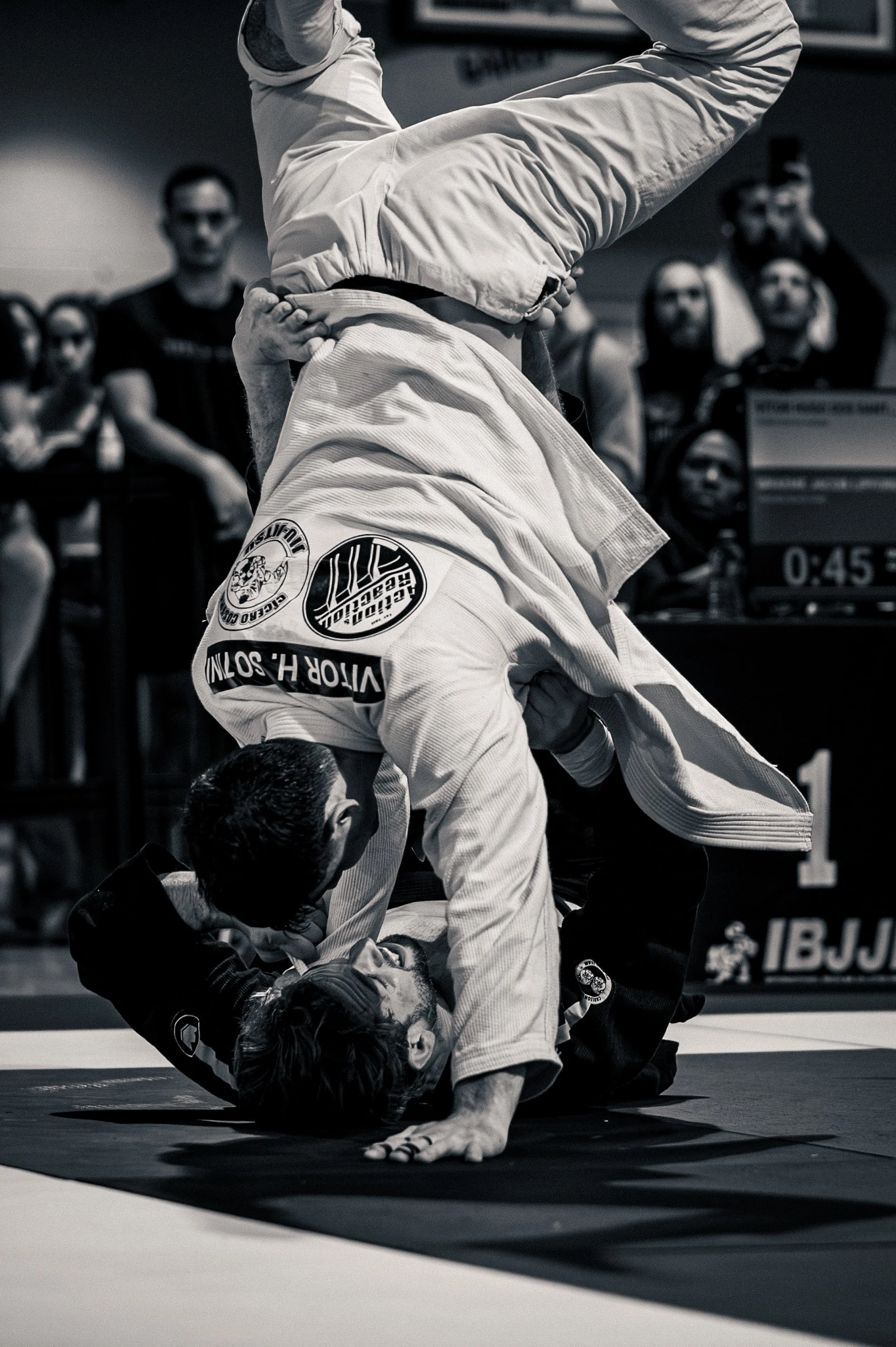 Two Brazilian Jiu-Jitsu practitioners in gis engaging in a grappling match on a mat, surrounded by spectators and an official in the background. Toronto Canada IBJJF