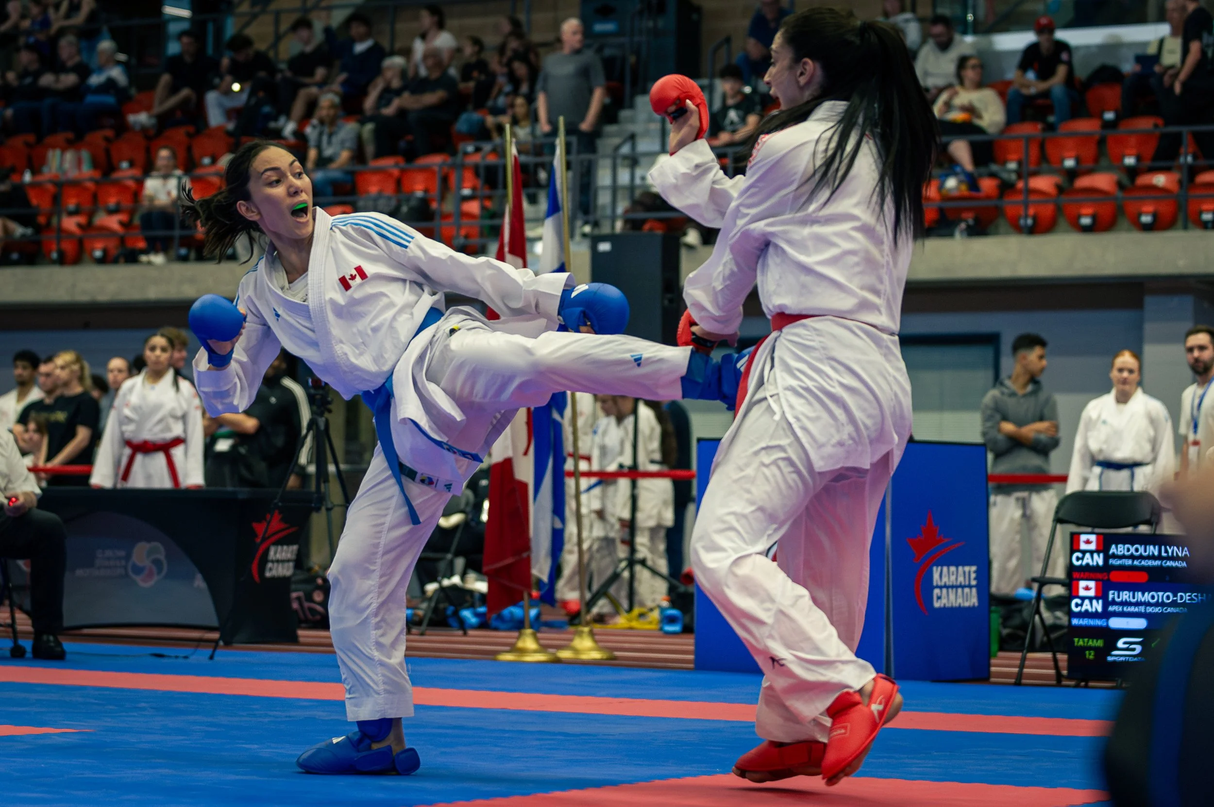 Two female martial artists compete in a karate match, one delivering a high kick to the other, in a sports arena with spectators in the background.