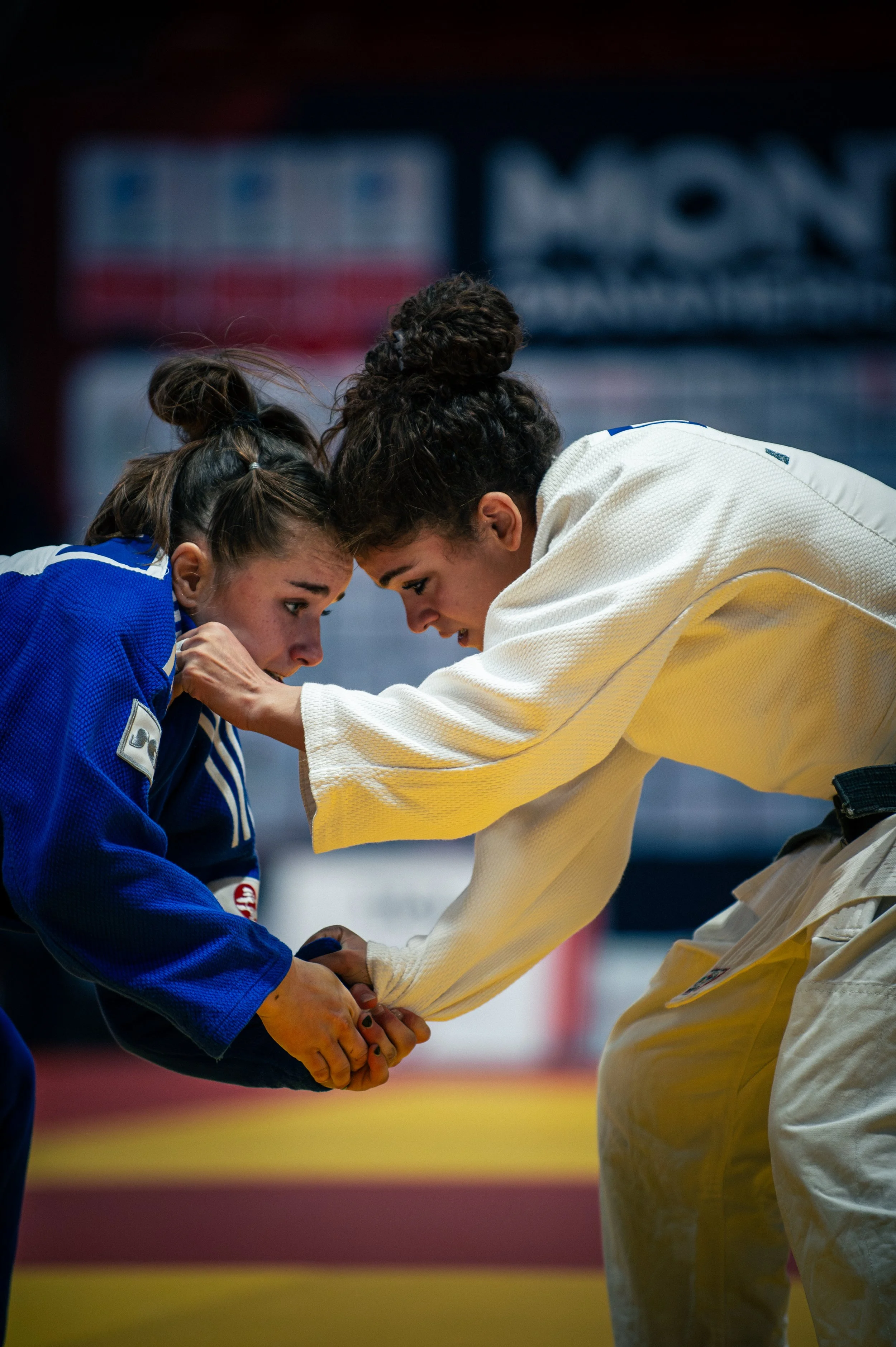 Two female judo competitors in a match, engaging in a handshake or grip, wearing blue and white judogis, on a training or competition mat.