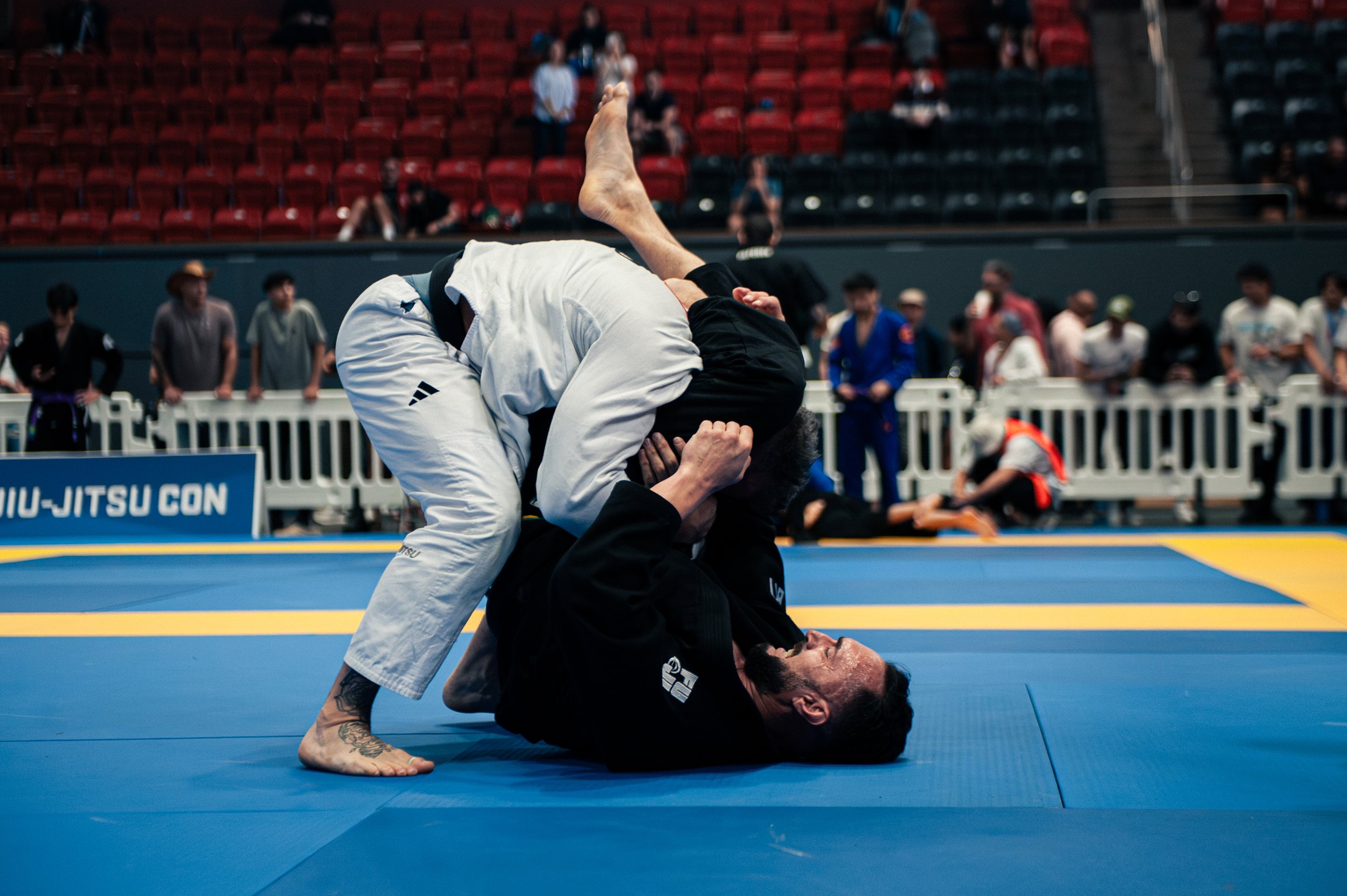 Two men practicing Brazilian Jiu-Jitsu on a blue mat in an indoor arena, with one man holding the other down while the other attempts to escape, with spectators in the background.