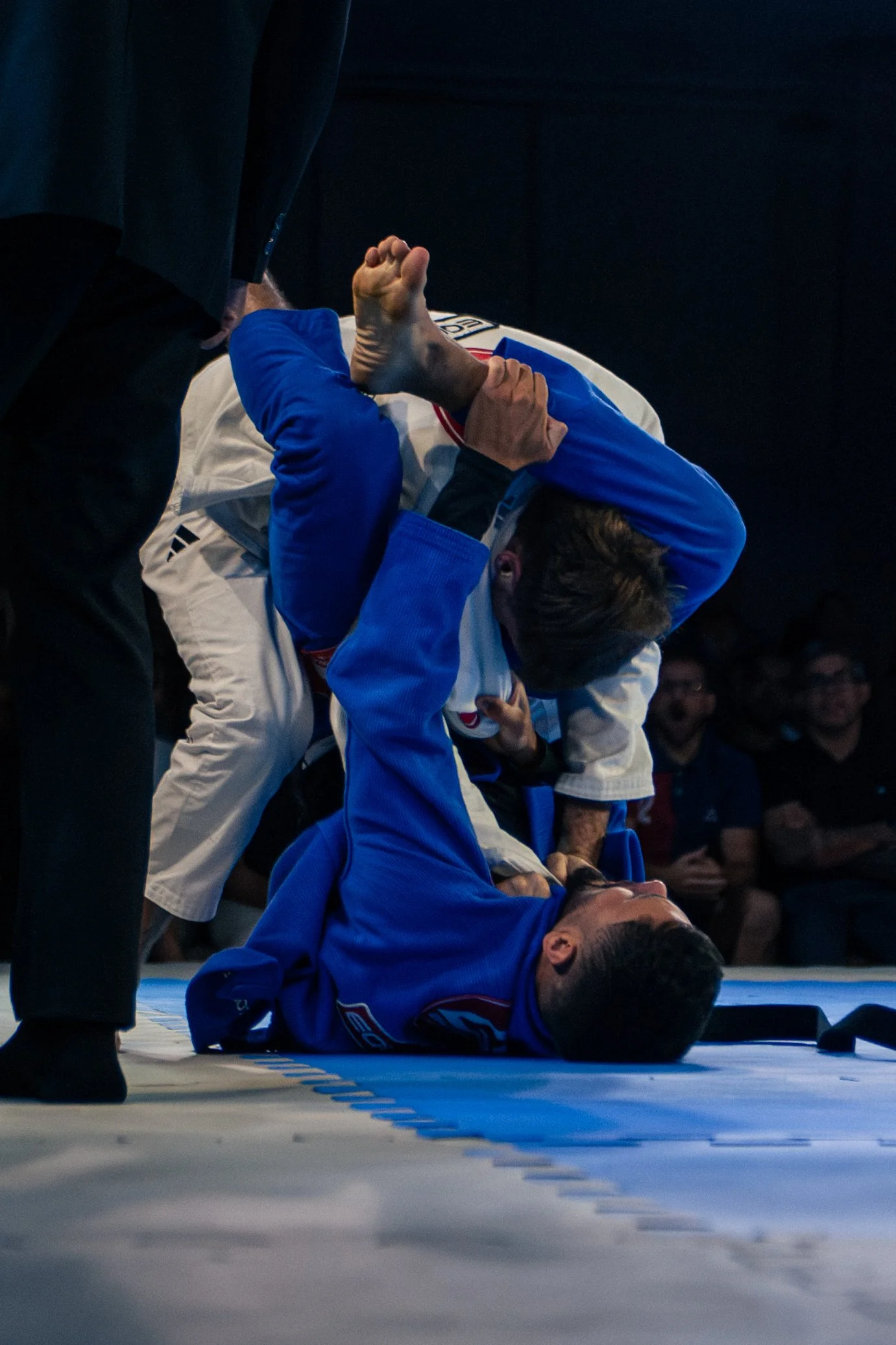 A martial arts competition with a male competitor in a blue gi lying on the mat, holding his head with his hand, while another competitor dressed in a white gi with a black belt is standing nearby. An official in black pants is also visible.