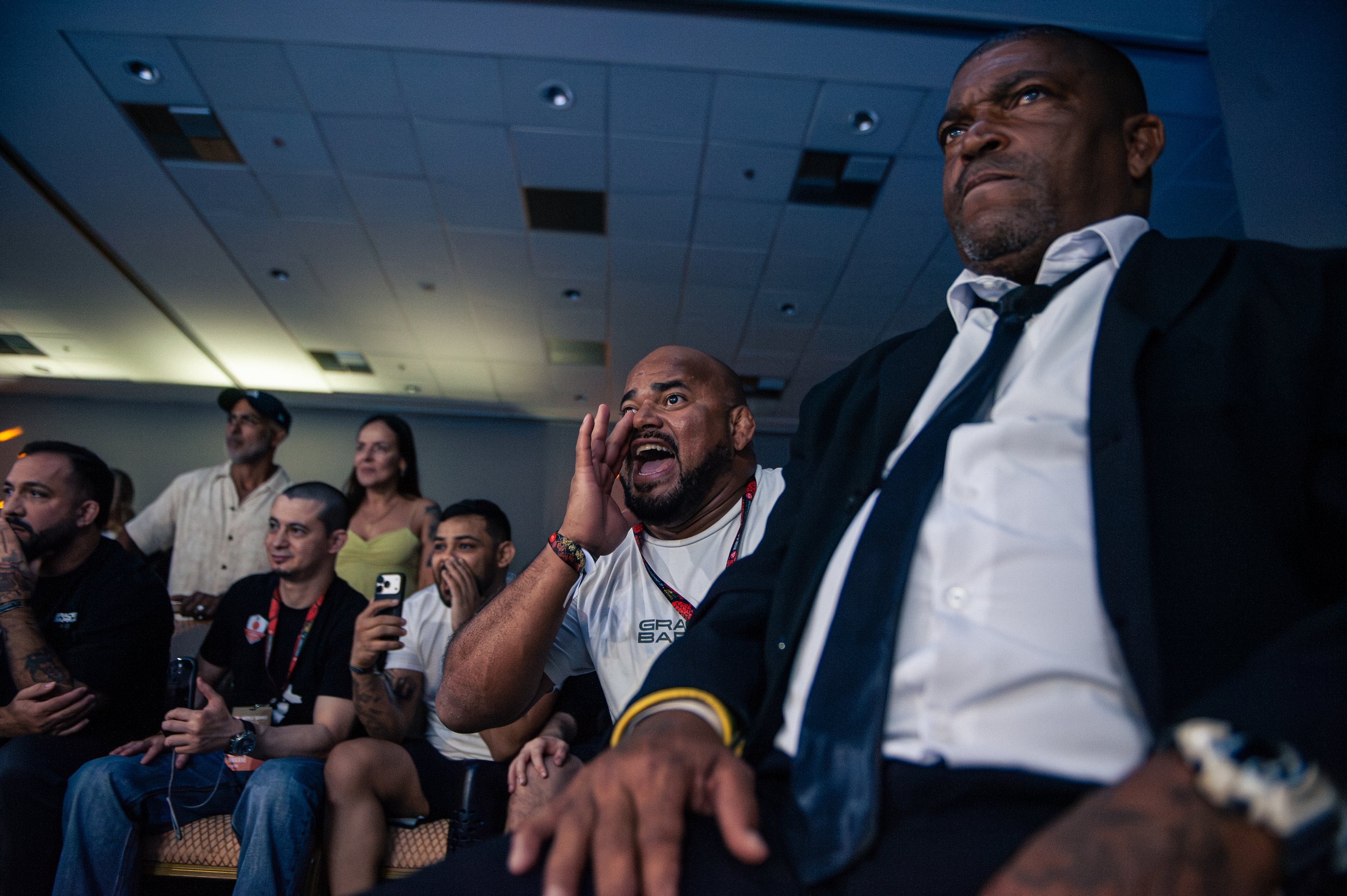 A group of diverse people at Gracie Barra Super Fights, some are reacting excitedly or surprised, with a man in a suit in the foreground and others in casual attire behind him, in Rio.