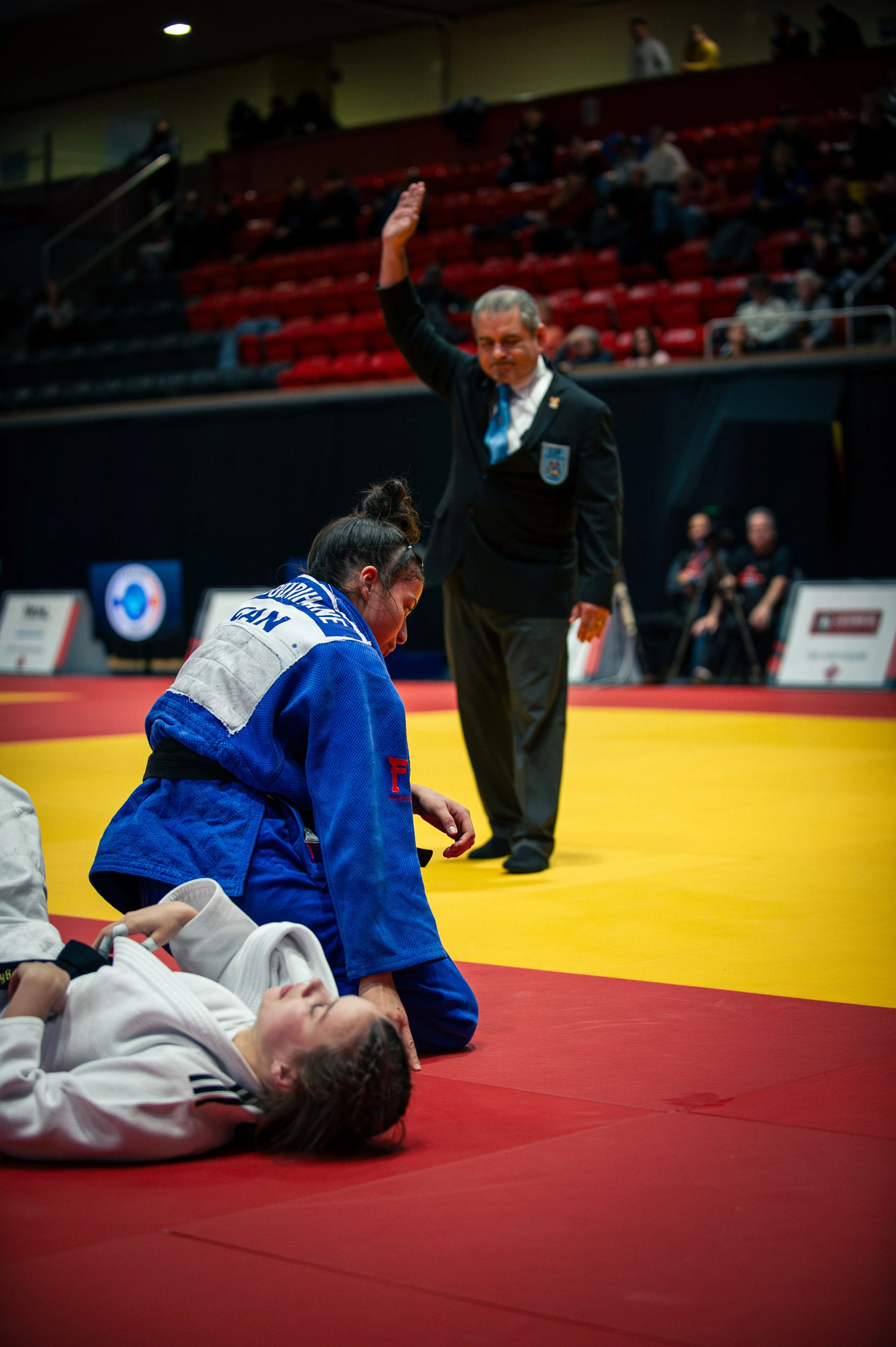 A female judo competitor in a blue gi receives medical attention on the mat, while a male referee in a suit raises his hand in a gesture during a match. There are spectators in the background. IJF Judo Canada
