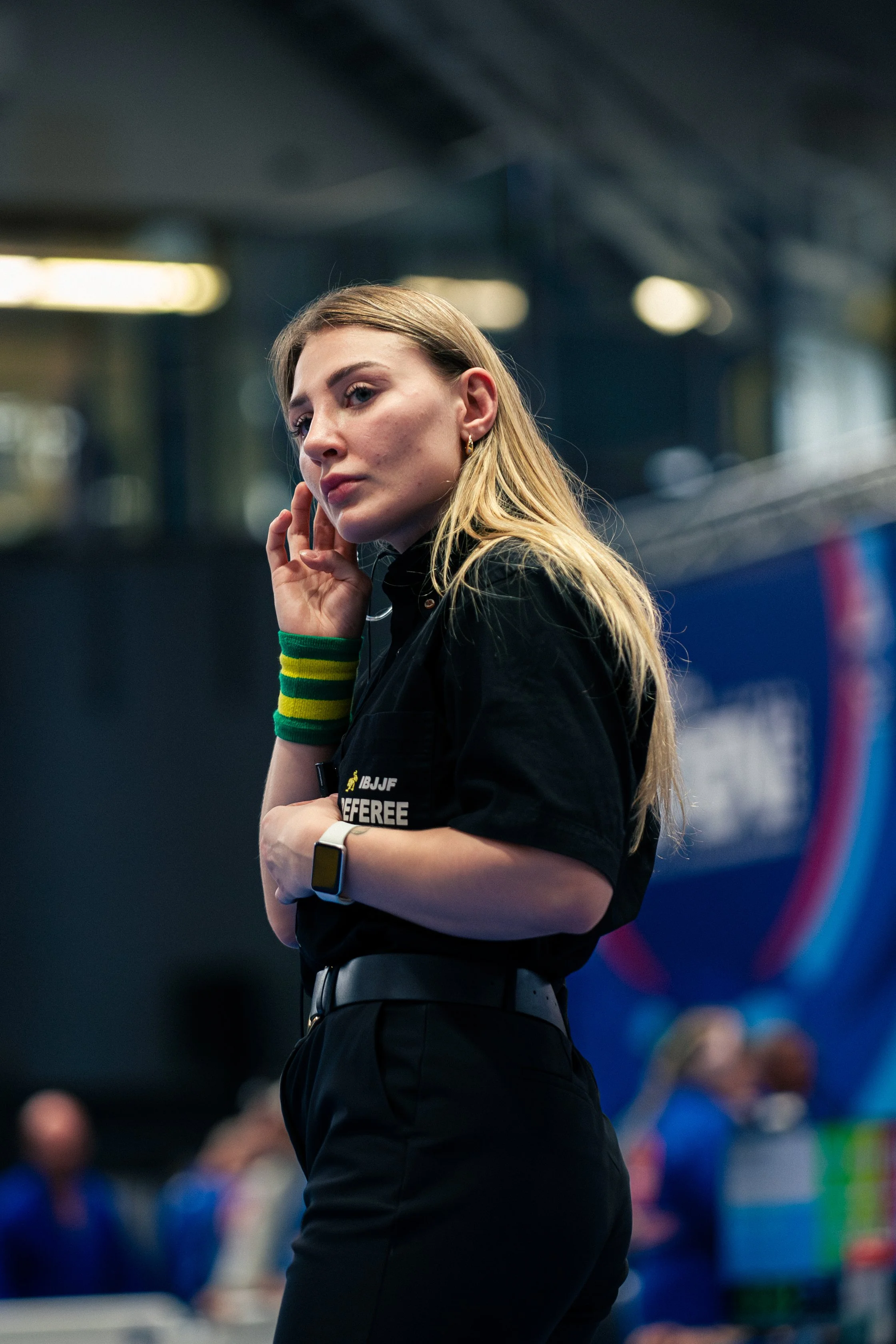 A woman with long blonde hair and a serious expression, wearing a black referee shirt with the logo IBJJF and the word 'Referee,' standing in an indoor sports arena.