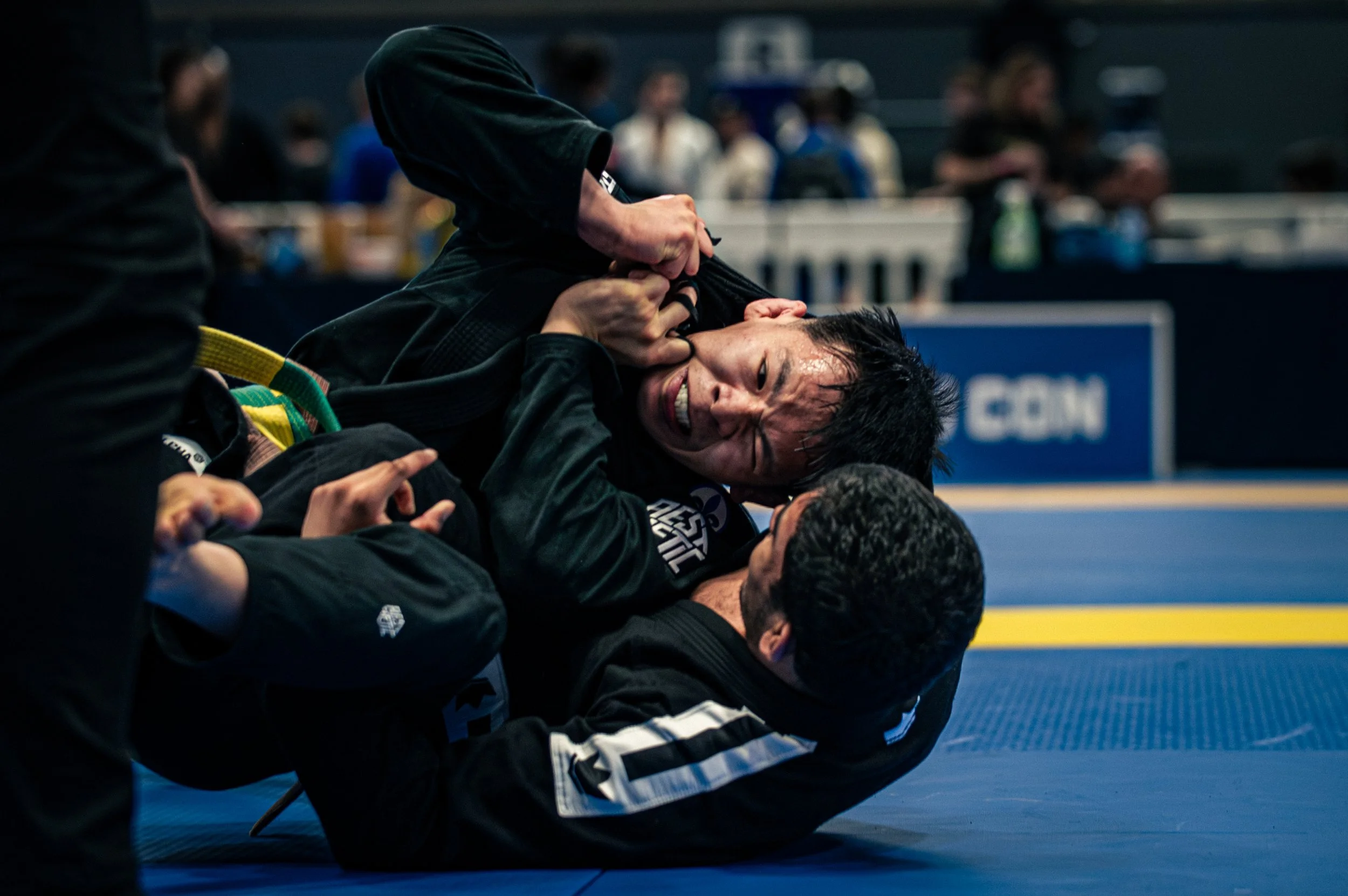 Two Brazilian Jiu-Jitsu practitioners in black gis engaged in a grappling match on a blue mat, with one on top of the other, in an indoor tournament setting with spectators blurred in the background. IBJJF Montreal