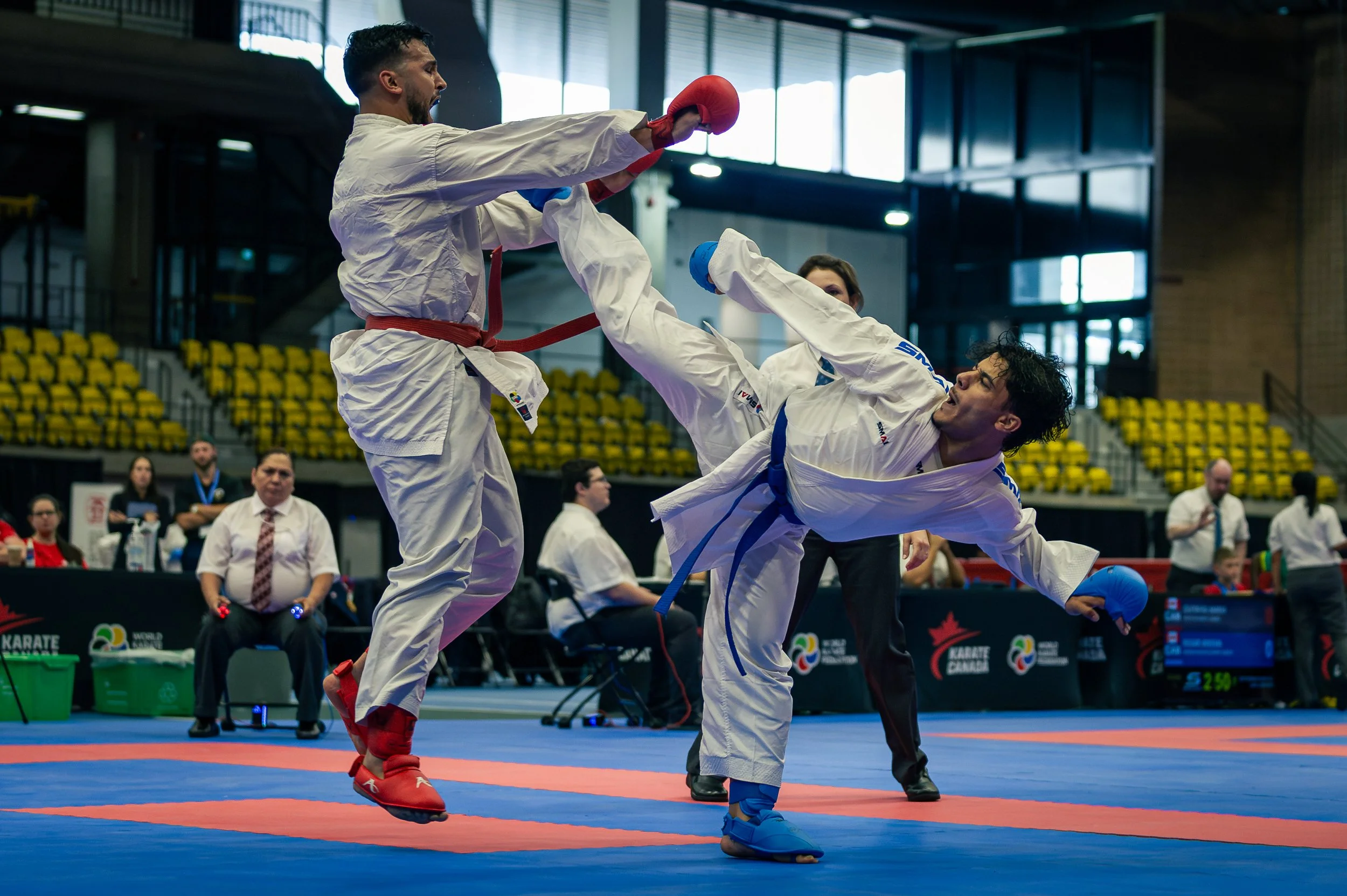 Two martial artists with protective gear engaged in a sparring match during a karate competition in an indoor sports arena. Judges and spectators are visible in the background.