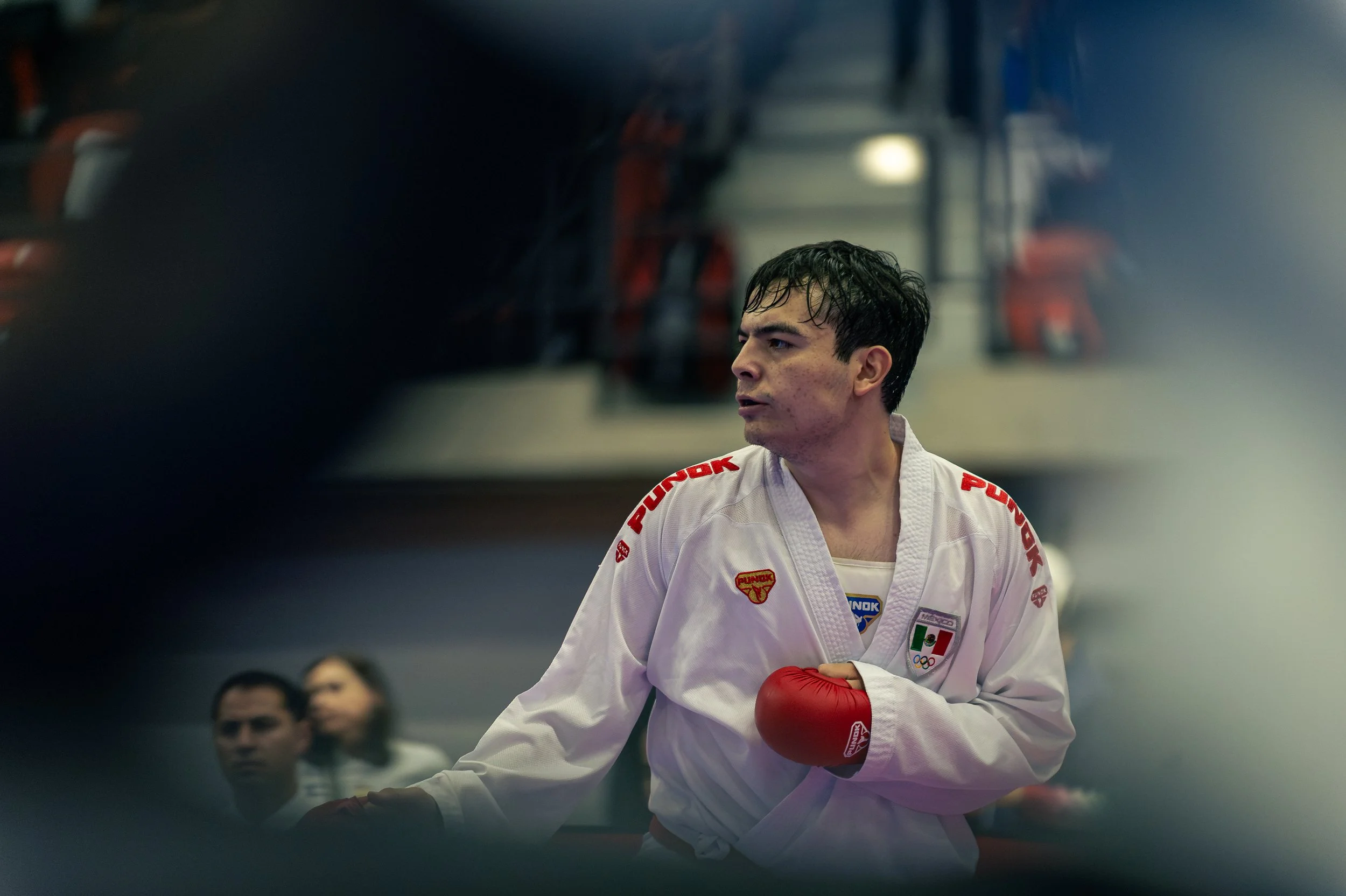 A male martial artist from mexico in a white gi with red gloves, wearing the Mexican flag and Olympic patches, competing in the montreal international open and people watching in the background.