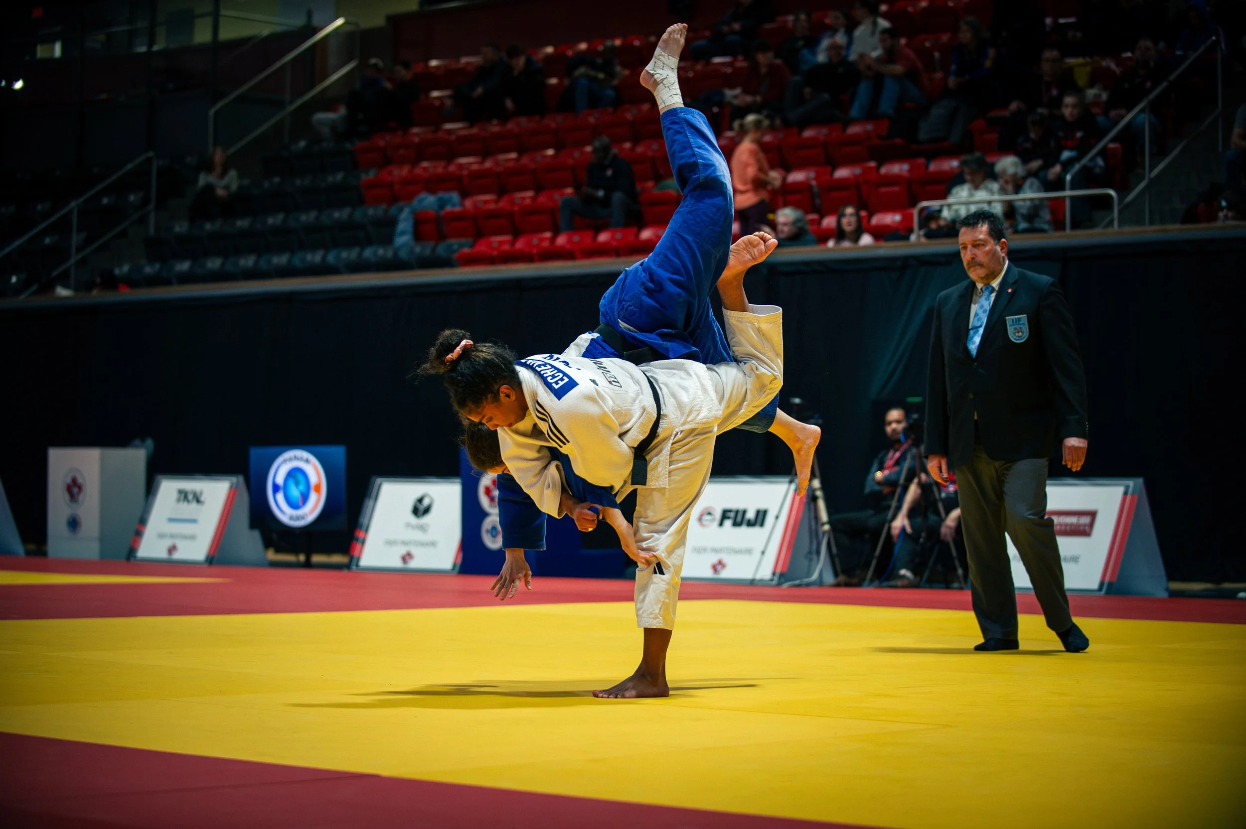 Two female judo competitors on a yellow mat, one performing a throw on the other, at a judo competition arena with judges and spectators in the background. IJF