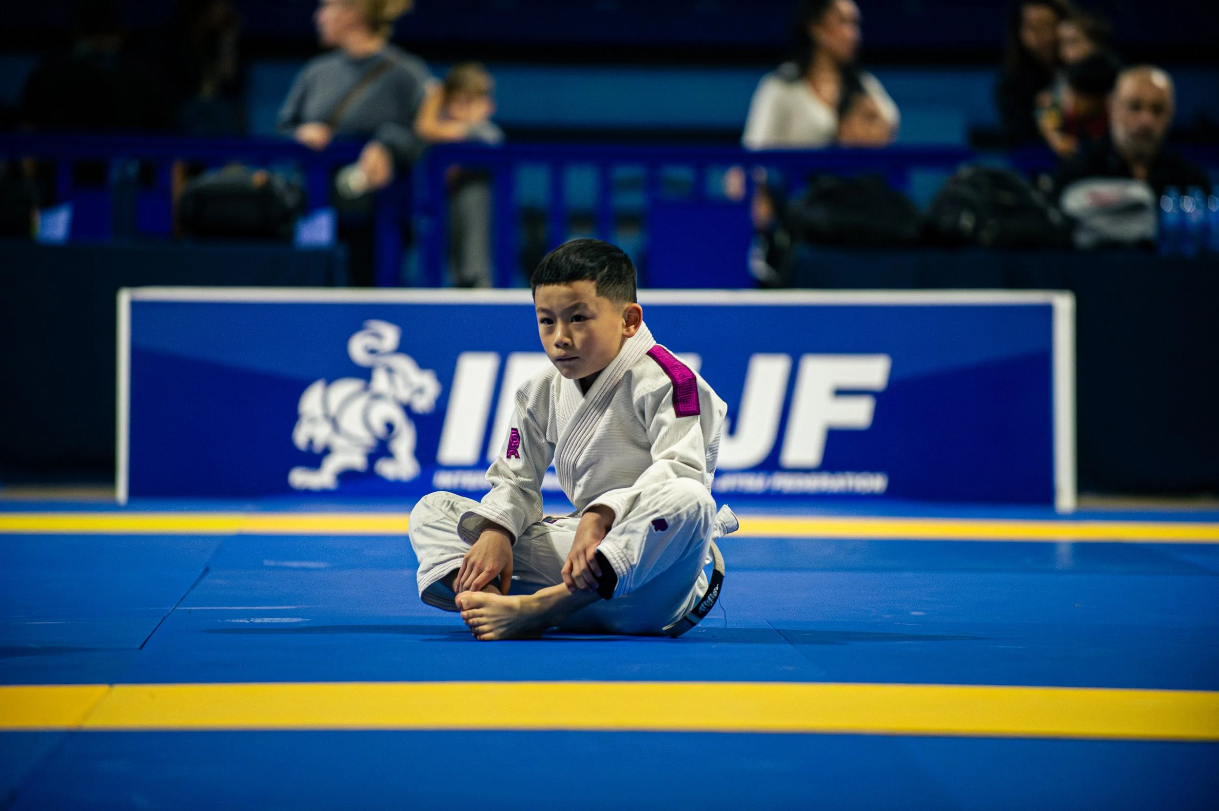 Young boy dressed in a martial arts uniform sitting on a blue and yellow mat, in a sports arena with spectators in the background. IBJJF
