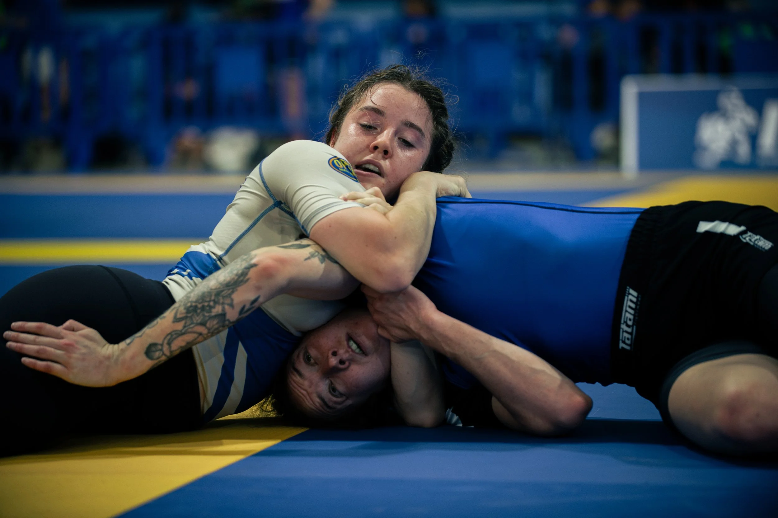 Two women competing in a Brazilian Jiu-Jitsu match on a blue mat, one woman in a white gi applying a submission hold on the woman in a black gi, both focused and intense.
