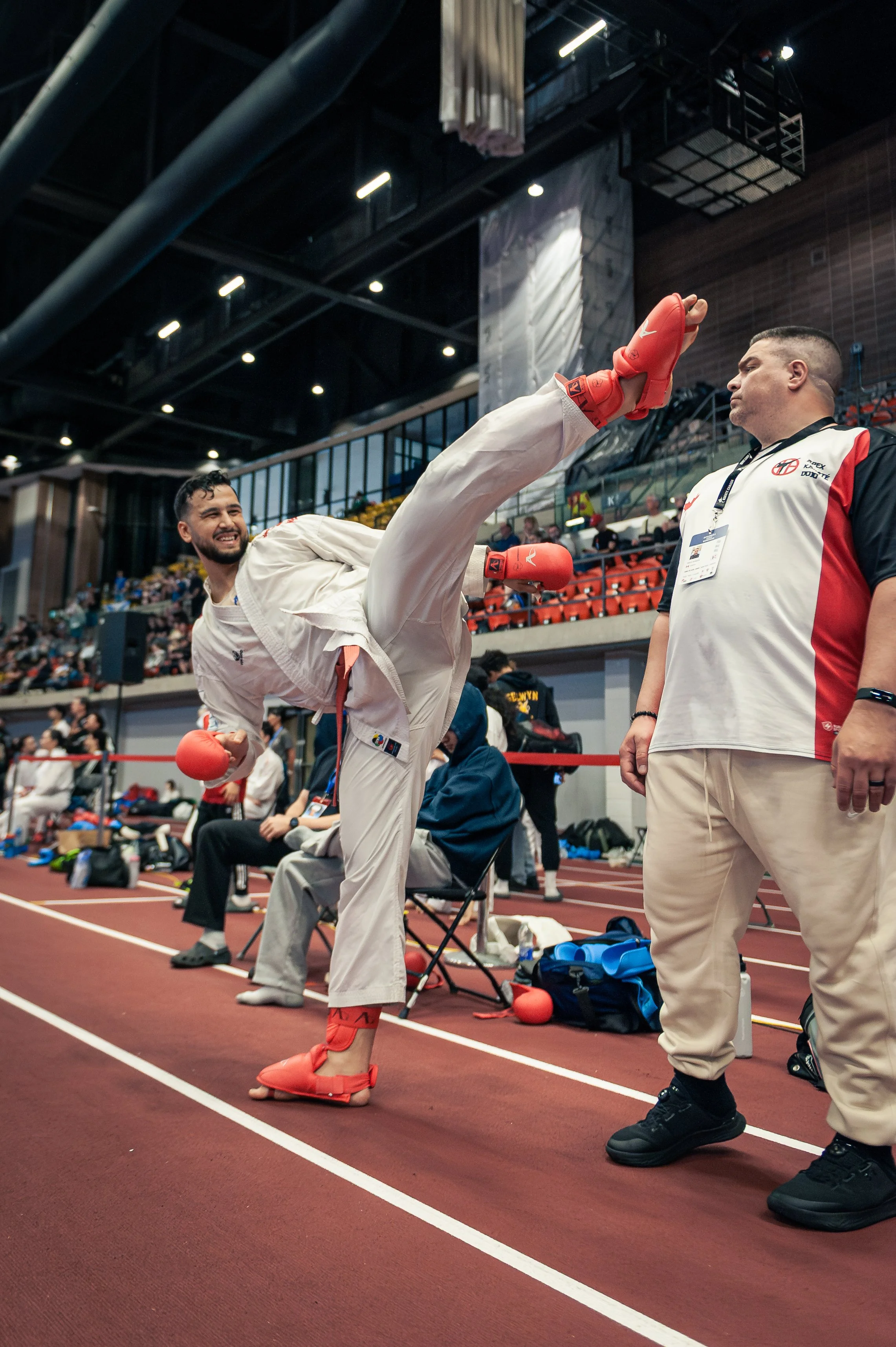 A male martial arts athlete in a white gi and red protective gear performing a high kick at an indoor sports arena during a competition, with a referee or coach observing nearby.