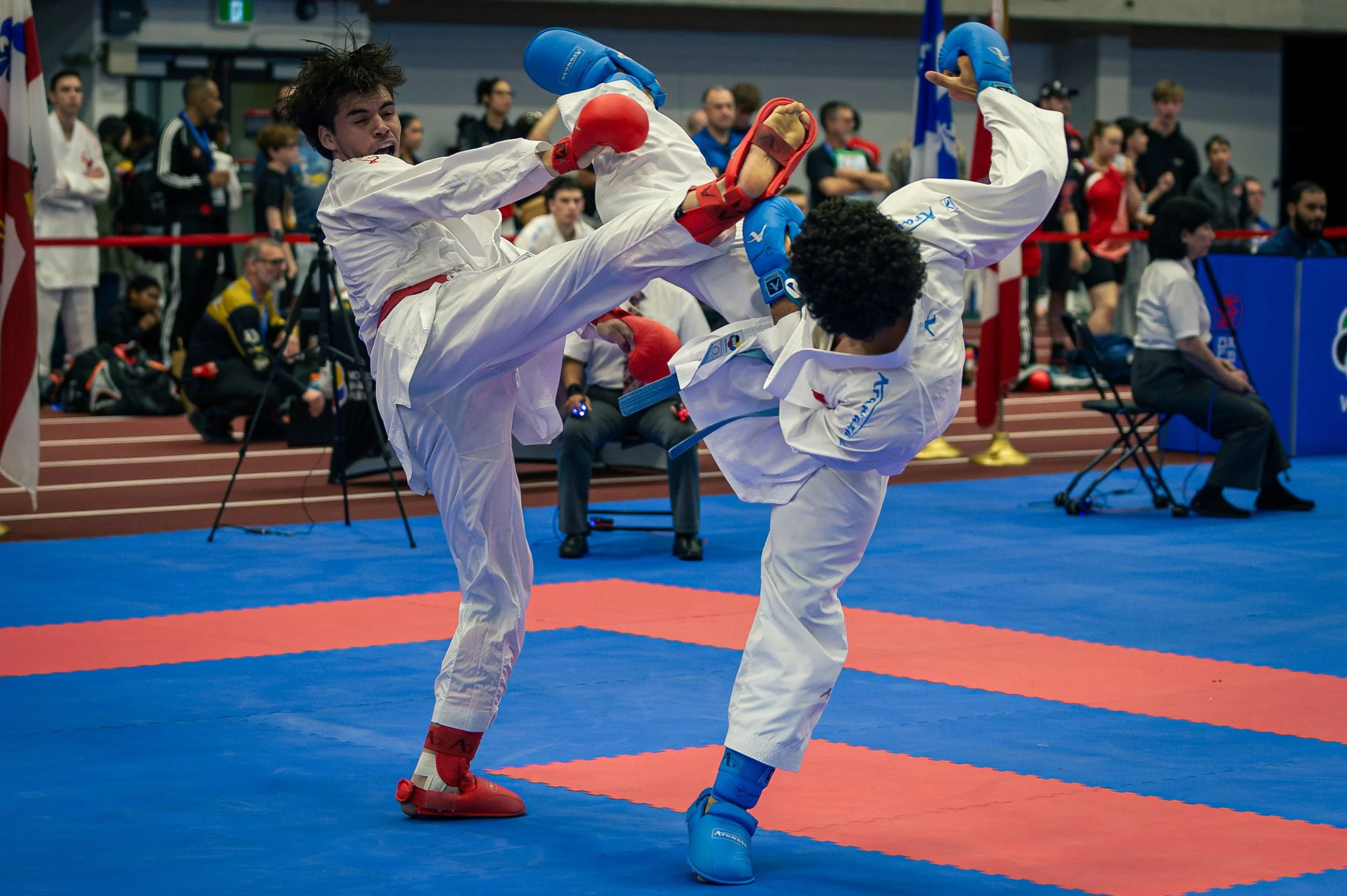 Two martial artists in white gi uniforms and protective gear sparring in a karate competition, with spectators watching in the background.