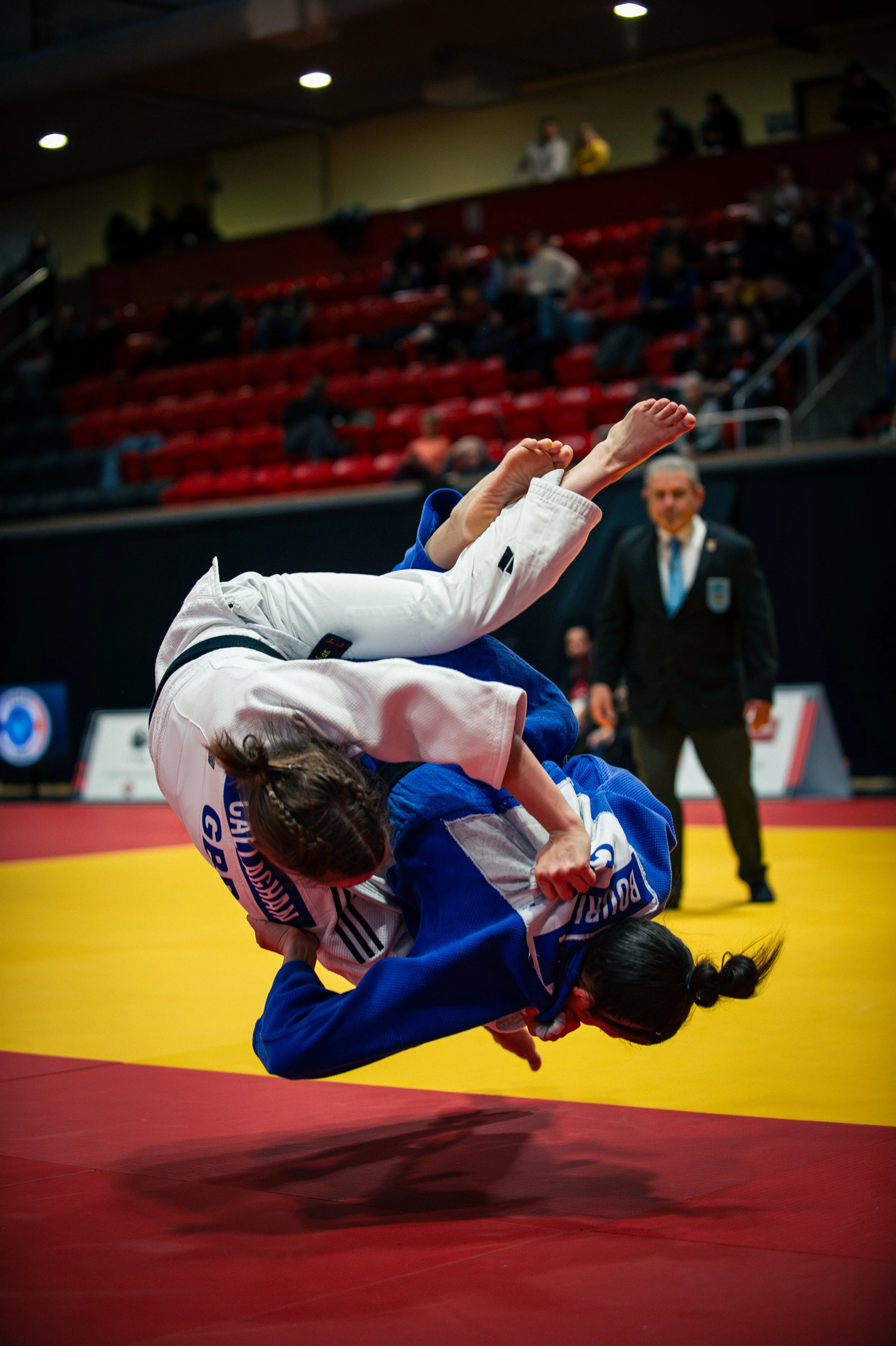 Two female judo competitors in judo uniforms are engaged in a match on a yellow and red mat, with one executing a throw on the other. An official in a suit watches in the background, and spectators are seated in the stands.