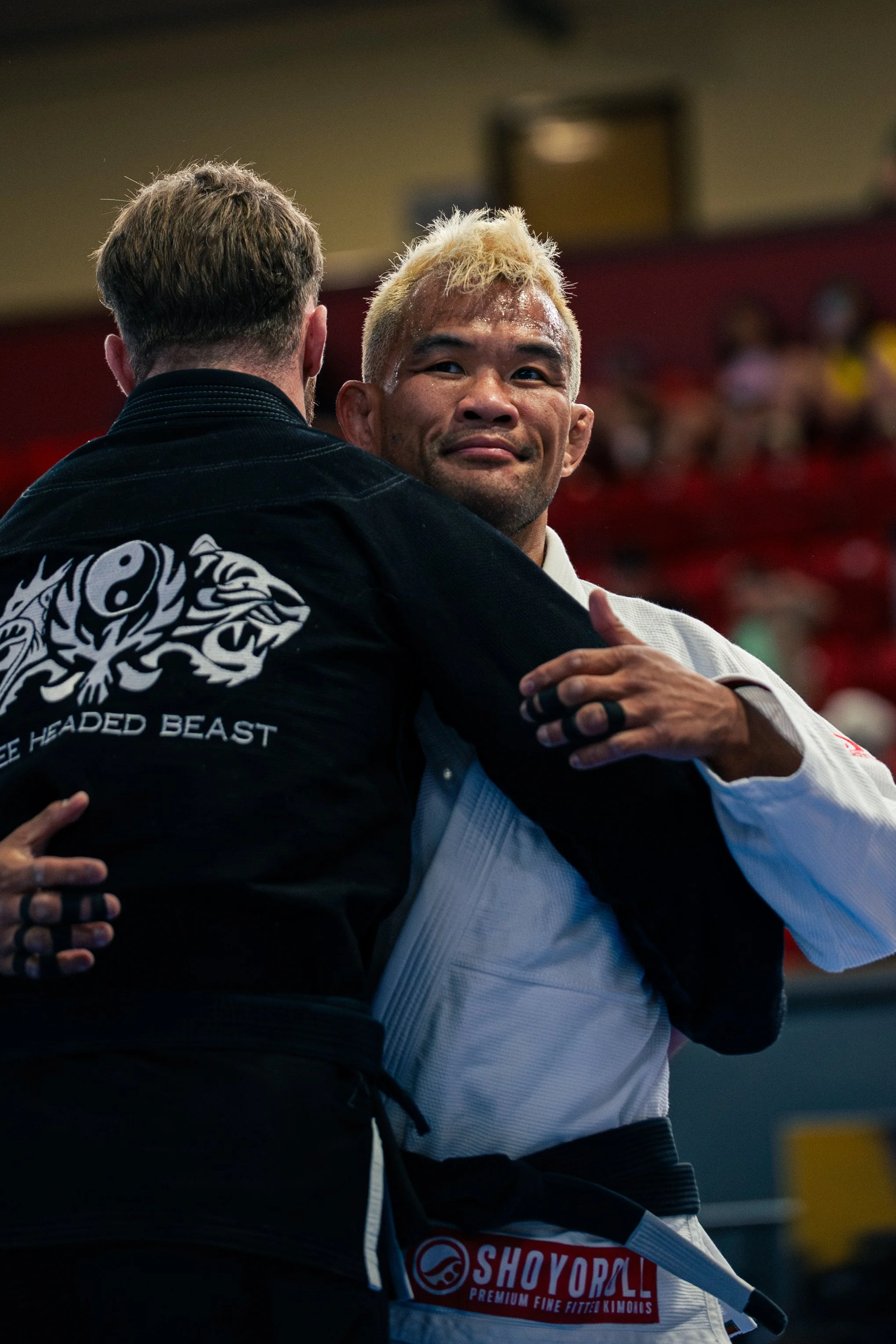 Two male martial artists, one with blonde hair and one with darker hair, embrace in a hug wearing martial arts uniforms, during a competition or event.