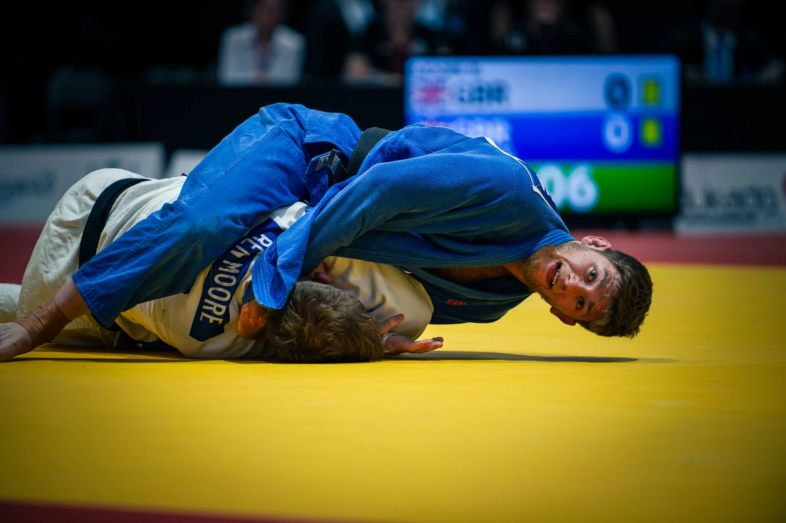 An intense moment in a judo match featuring one competitor in a white gi and another in a blue gi on a yellow mat, with a referee closely observing.