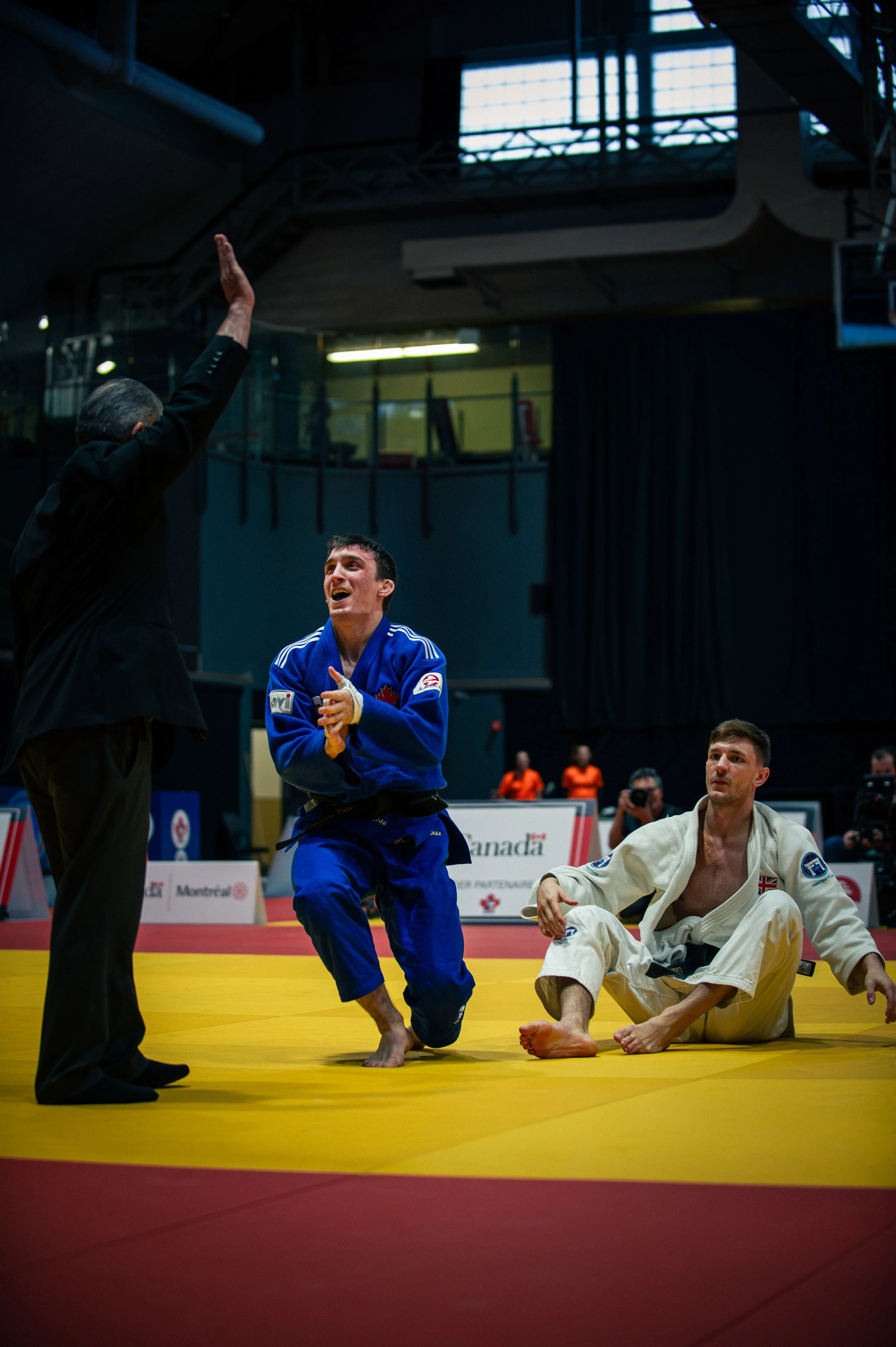 A match featuring two male competitors on a yellow mat. One fighter in a blue gi appears to be celebrating or expressing emotion, while the other in a white gi sits on the mat. An official in black officiates, and spectators are visible in the backgr