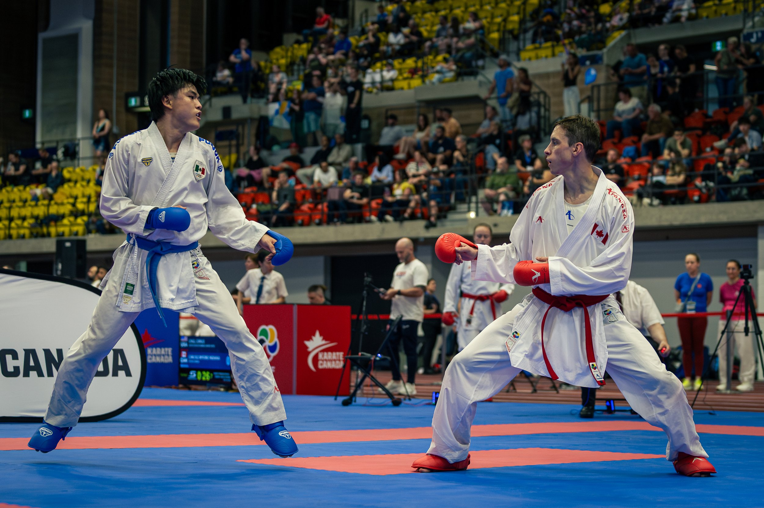 Two martial artists, one with a blue belt and gloves representing Mexico, and the other with a red belt and gloves representing Canada, competing at the montreal international open in a sports arena with spectators watching.