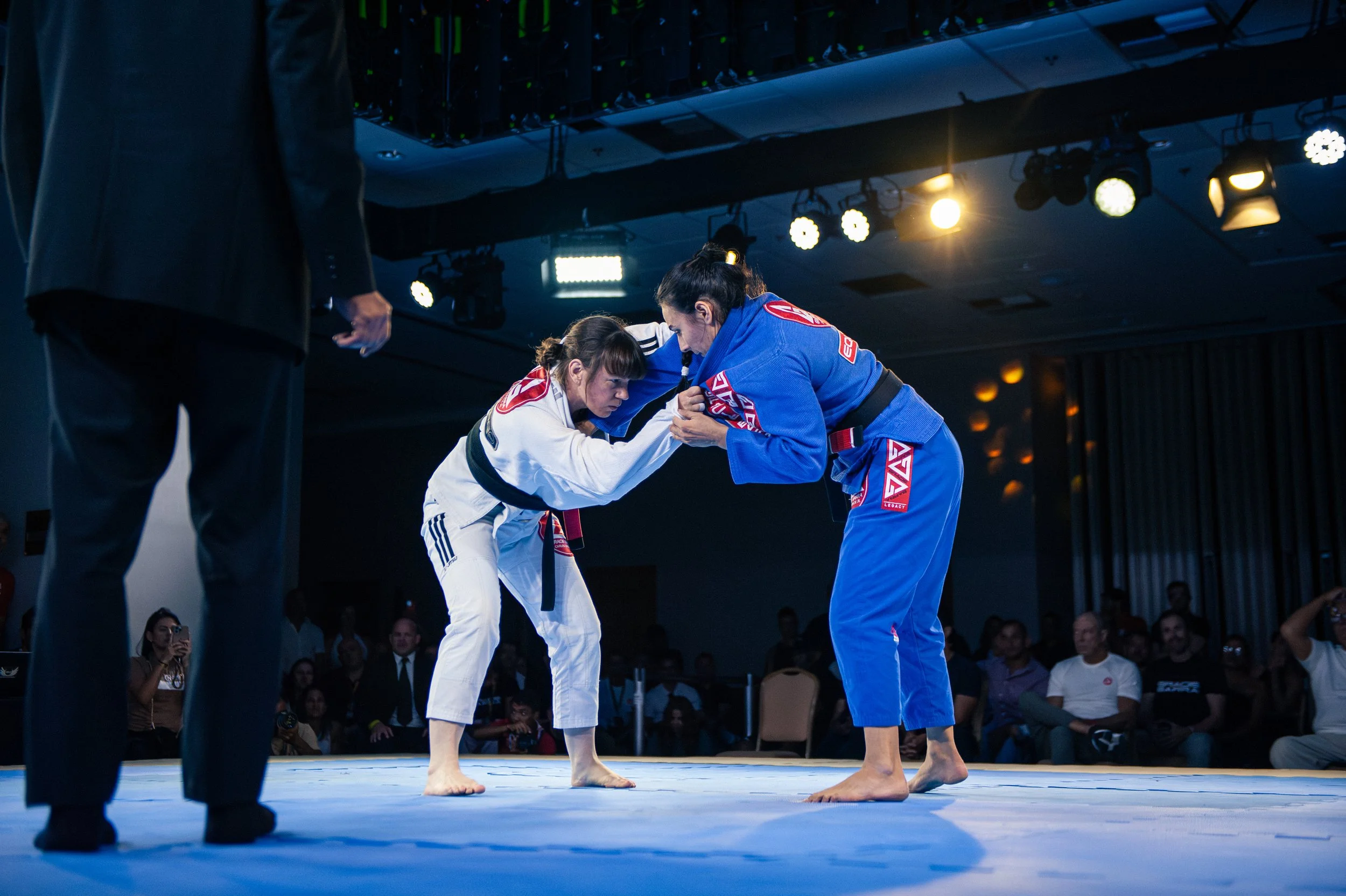Two female mixed martial artists, one in a white gi and the other in a blue gi, are engaged in a grappling match during a competition. They are facing each other with serious expressions, and an official in black is observing from the side. Audience 