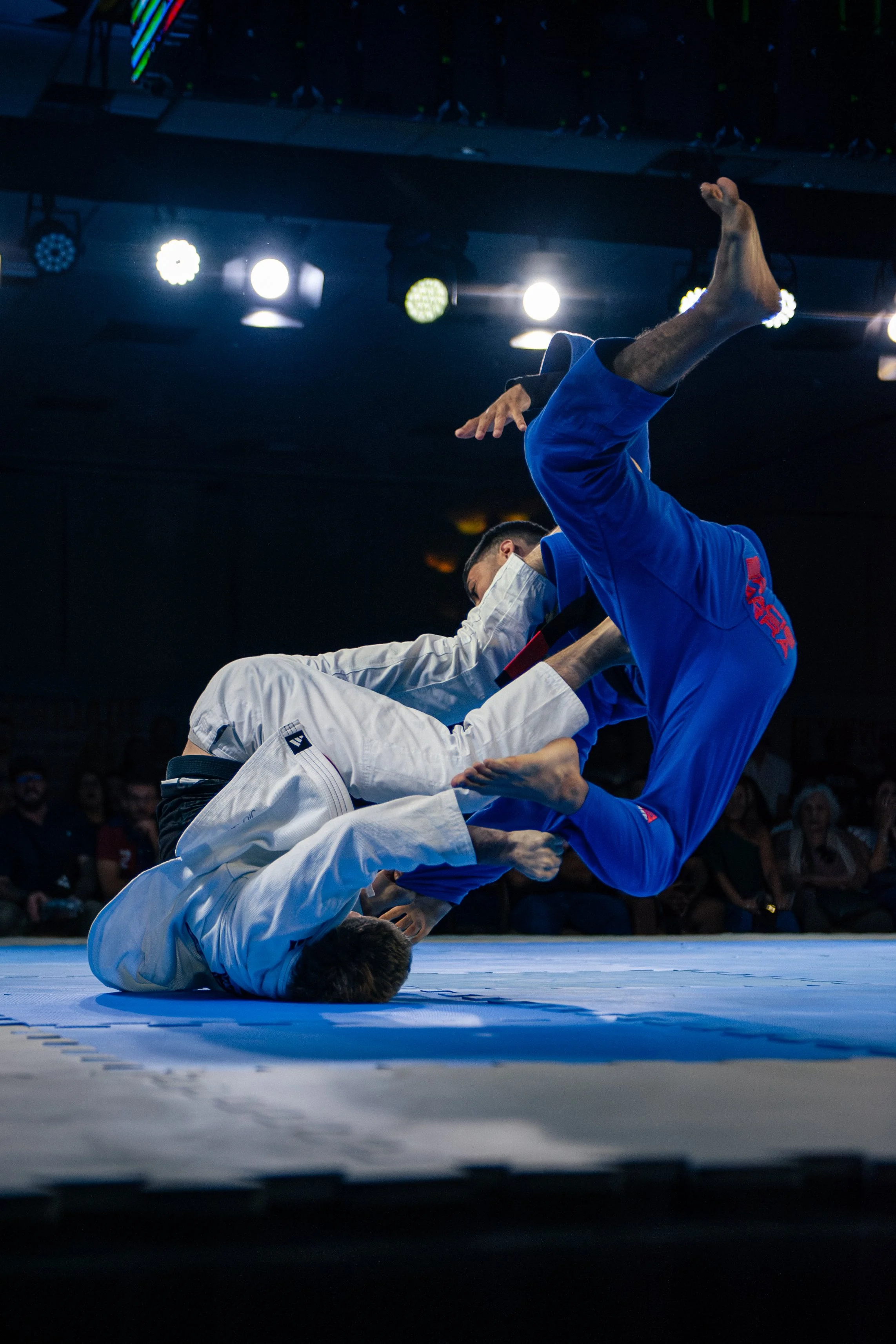 Two martial artists are practicing a technique on a blue mat in a gym. One is wearing a white gi, and the other is wearing a blue gi. They are in a wrestling position, with the person in white on the ground and the person in blue executing a move.
