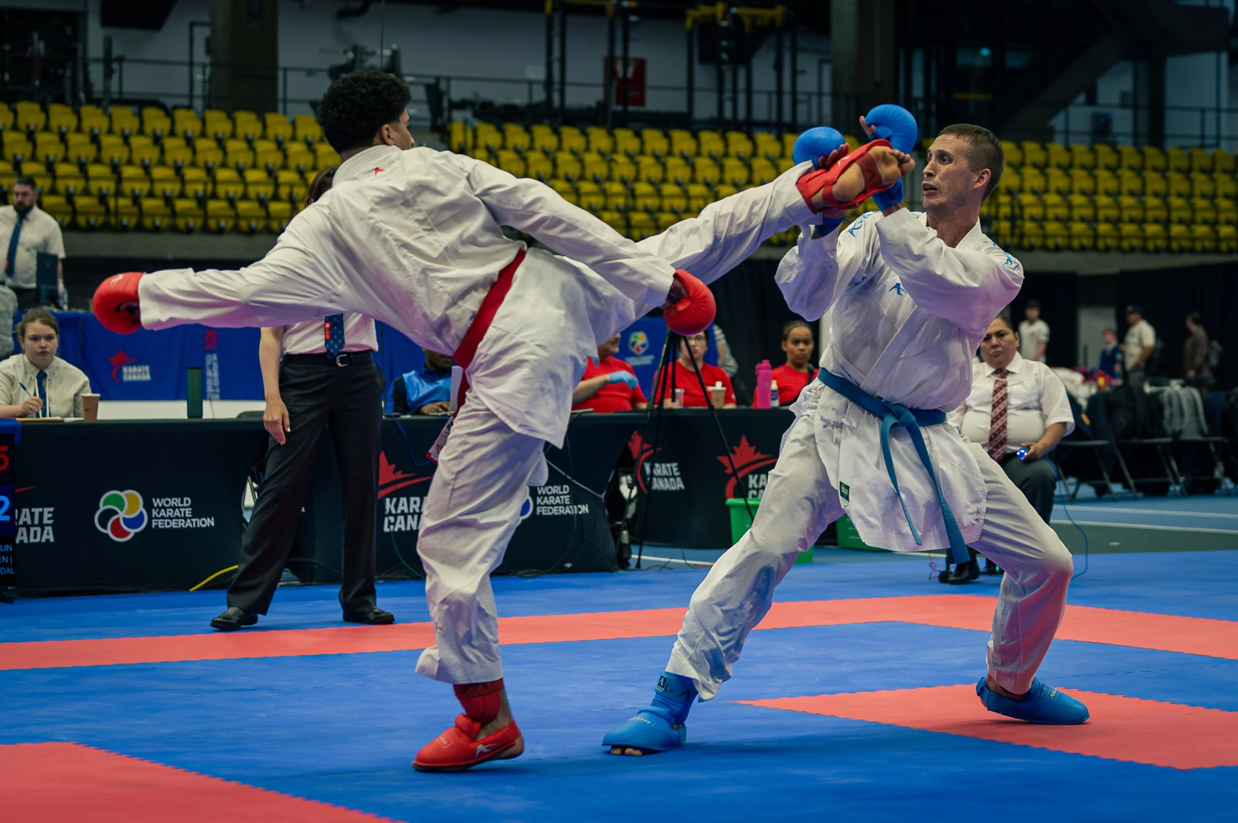 Two martial artists in white uniforms are sparring on a red and blue mat at a karate competition, with one delivering a high kick towards the other's face. Spectators and officials are visible in the background. montreal international open