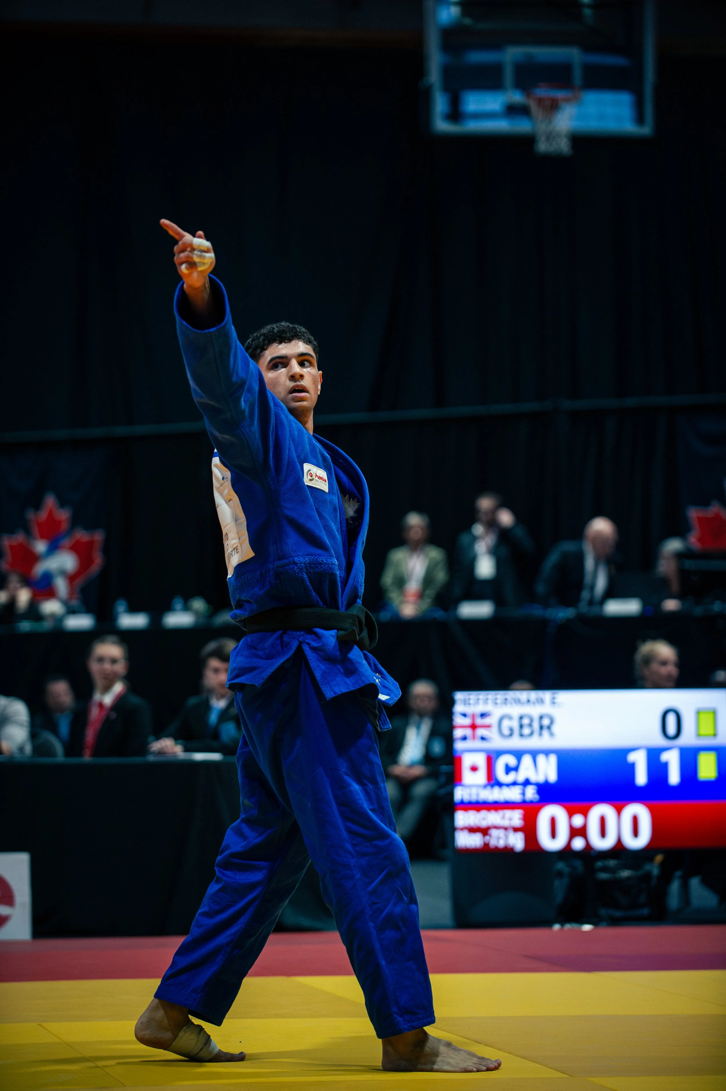 A male judo competitor in a blue gi, standing on a yellow mat, pointing with his right hand during a match. There is a scoreboard in the background showing the scores for GBR and CAN teams.