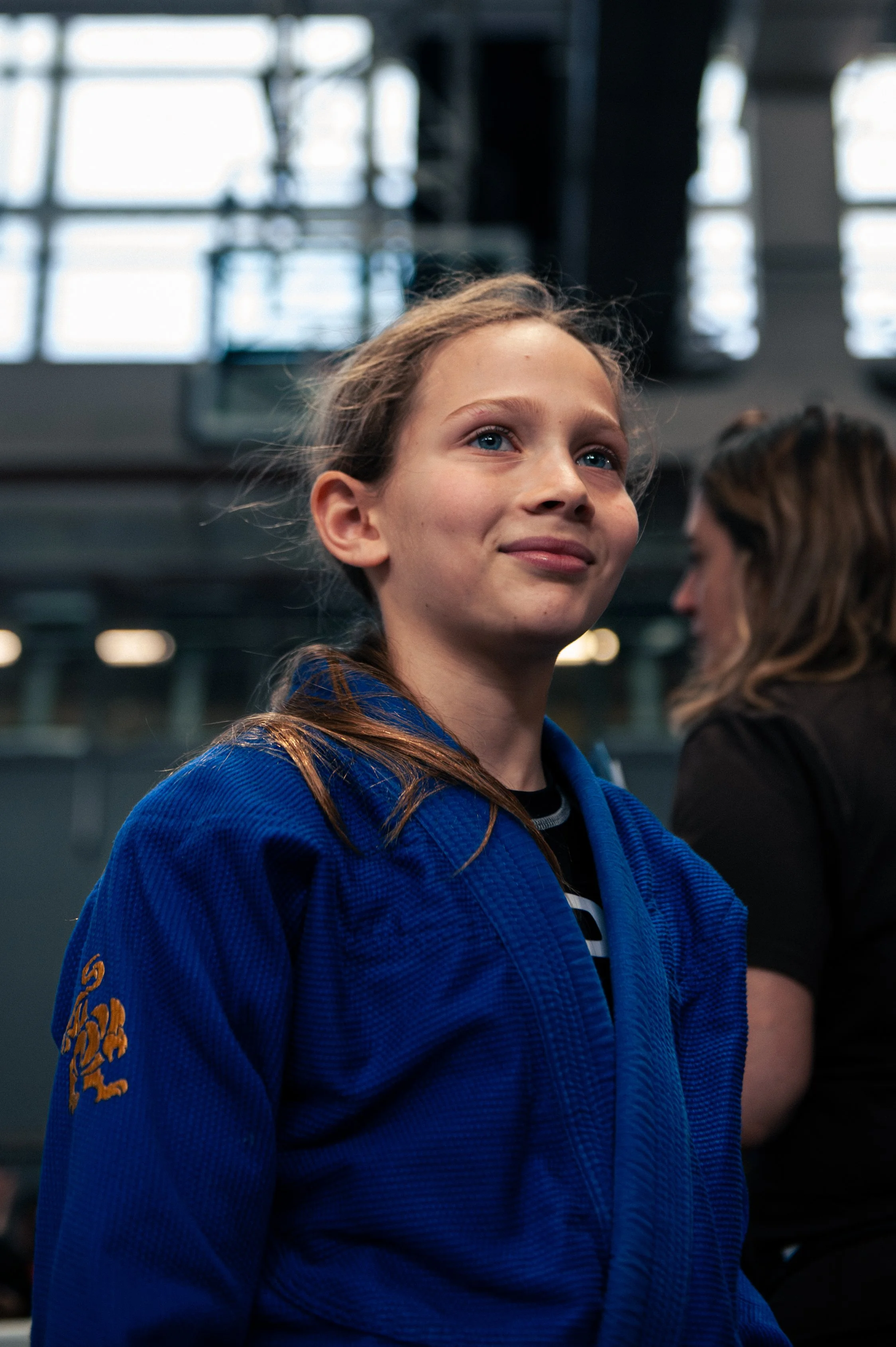 A young girl with blonde hair and blue eyes, wearing a blue martial arts uniform, standing indoors with a confident expression.
