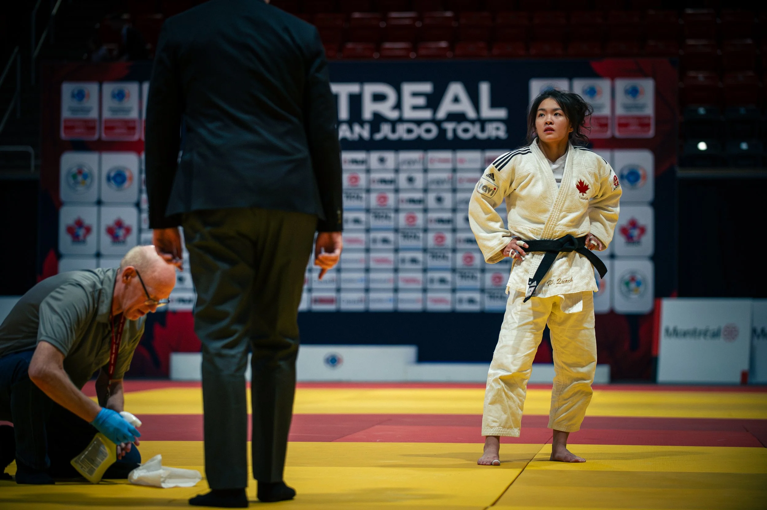 Young woman in a judo uniform standing on a tatami mat during a competition, with officials nearby, at MONTREAL JUDO TOUR event. IJF