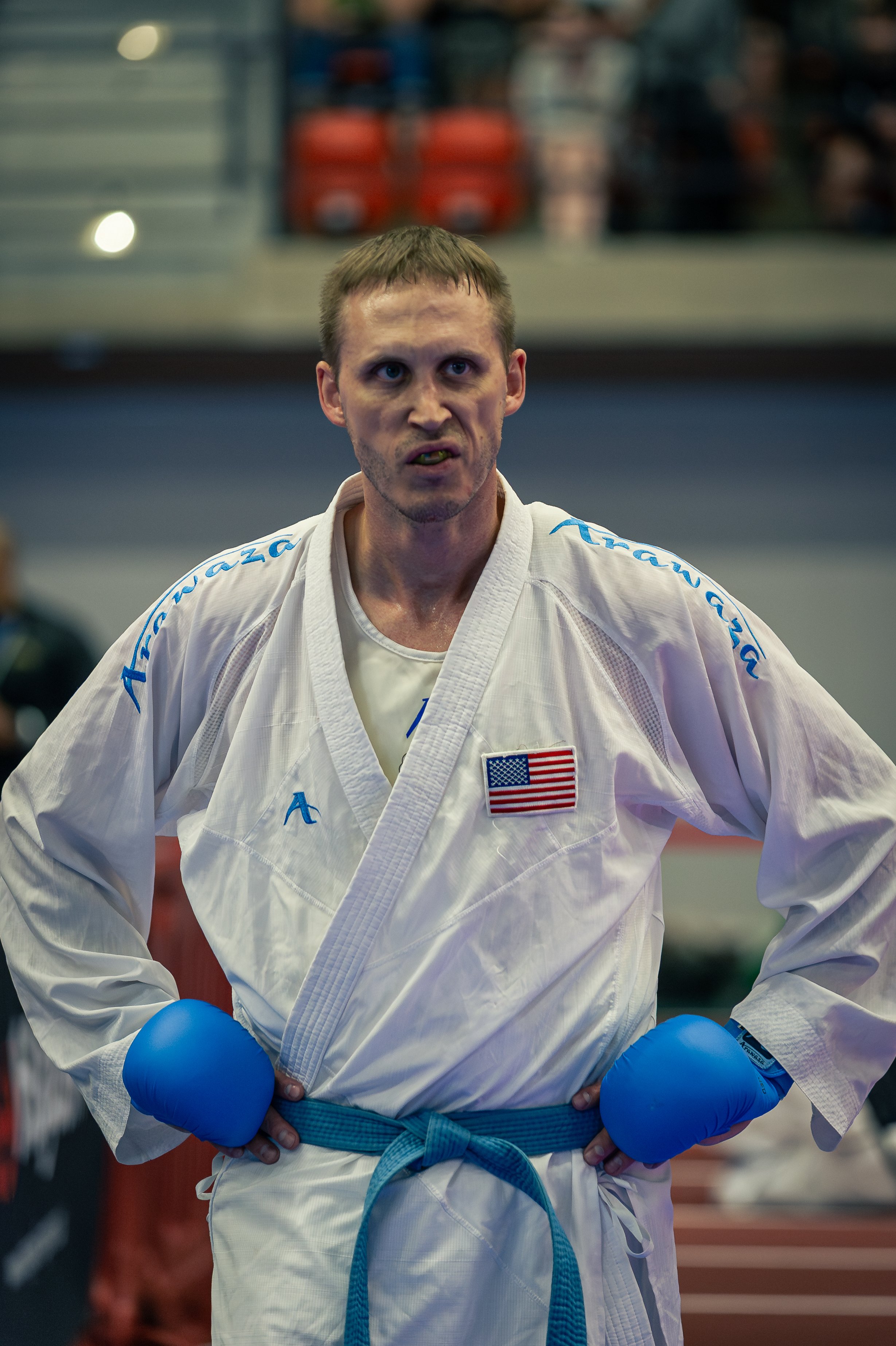 A male martial artist in a white gi with an American flag patch, blue gloves, and a blue belt, standing with his hands on his hips in an indoor sports facility.