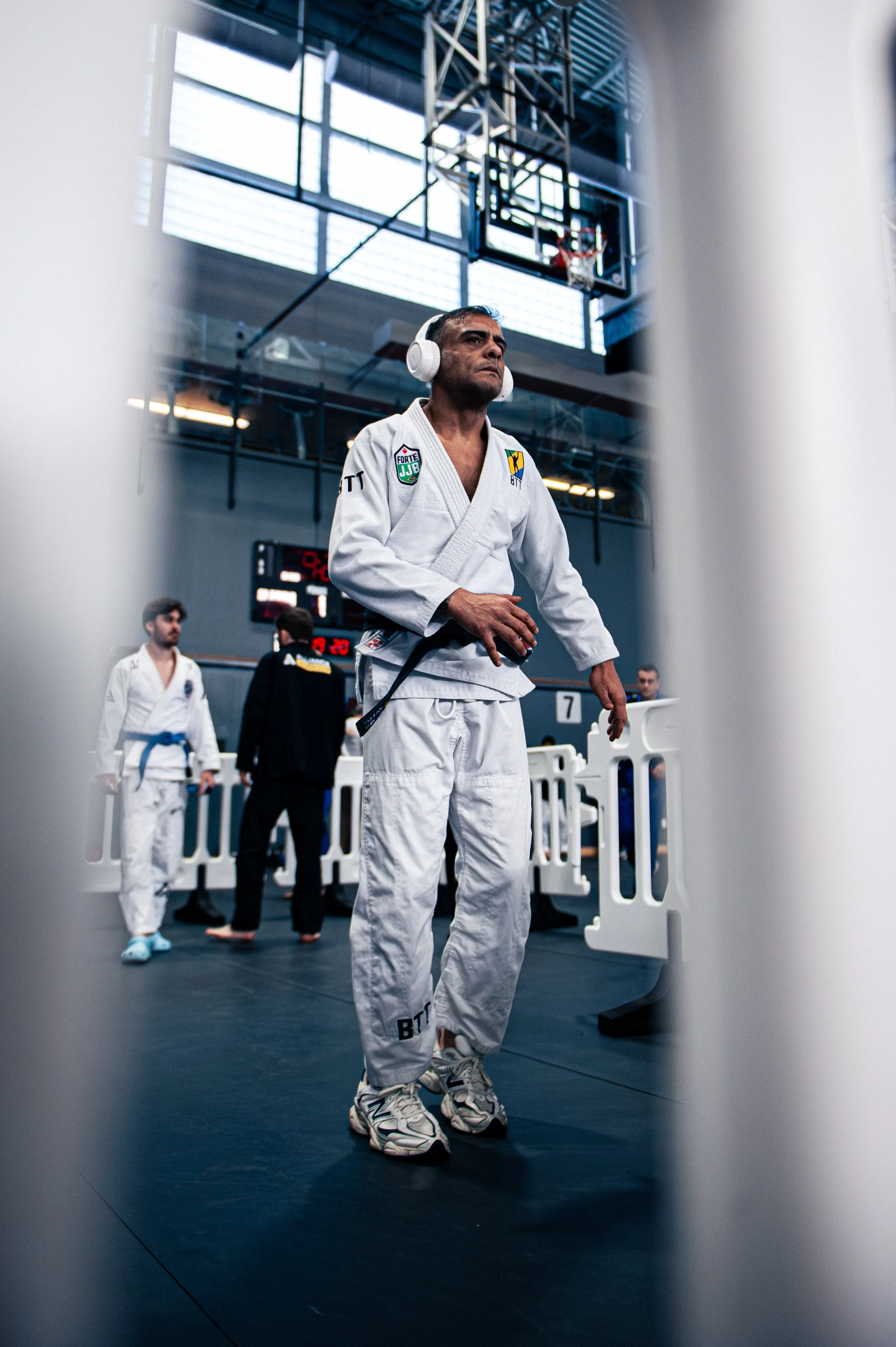 A man in a white BJJ gi with headphones standing in a gym, with others in gis and a basketball hoop in the background.