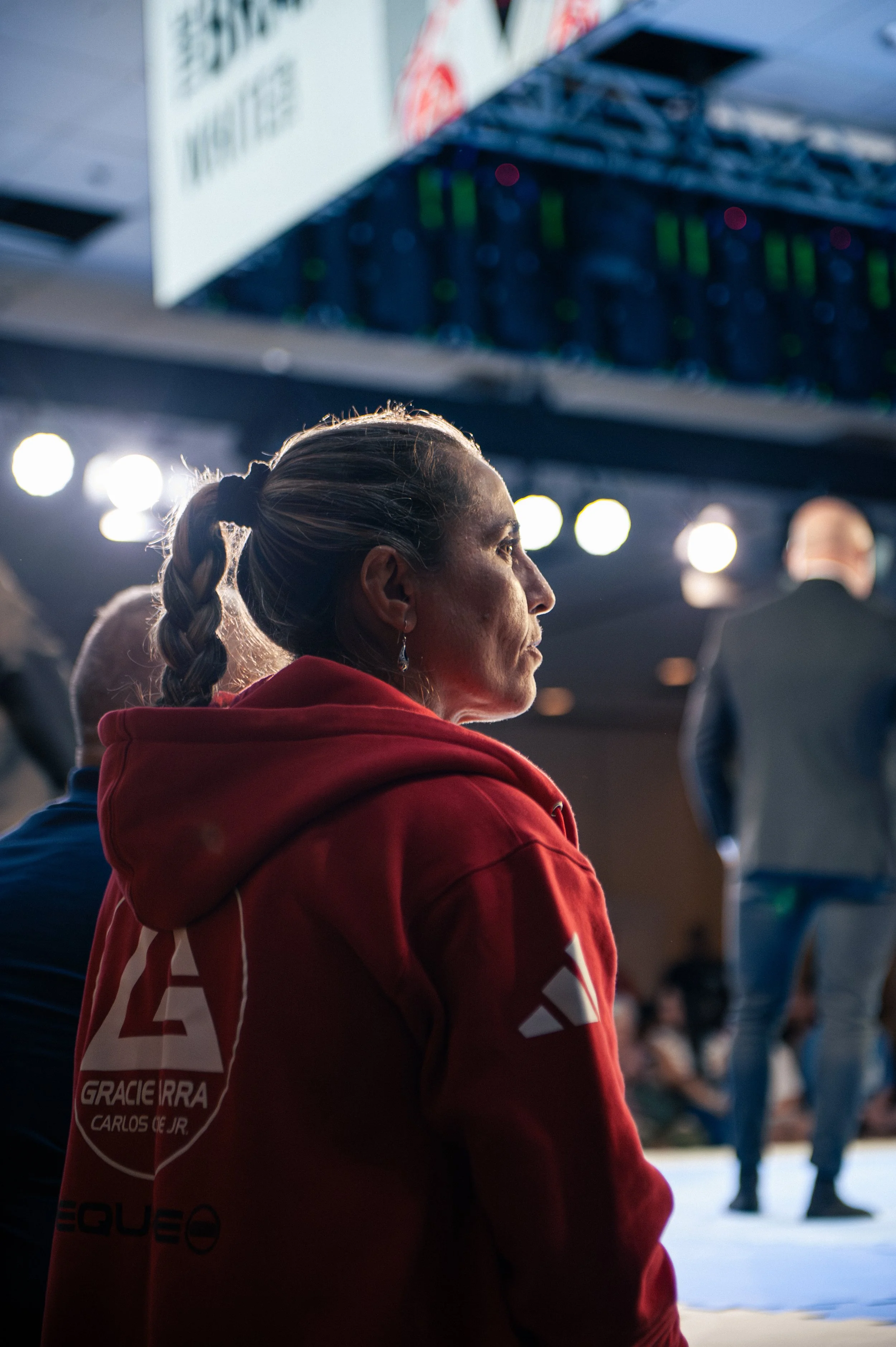 A woman with braided hair wearing a red hoodie, sitting in an indoor sports facility, possibly a gym or arena, with people and bright lights visible in the background.
