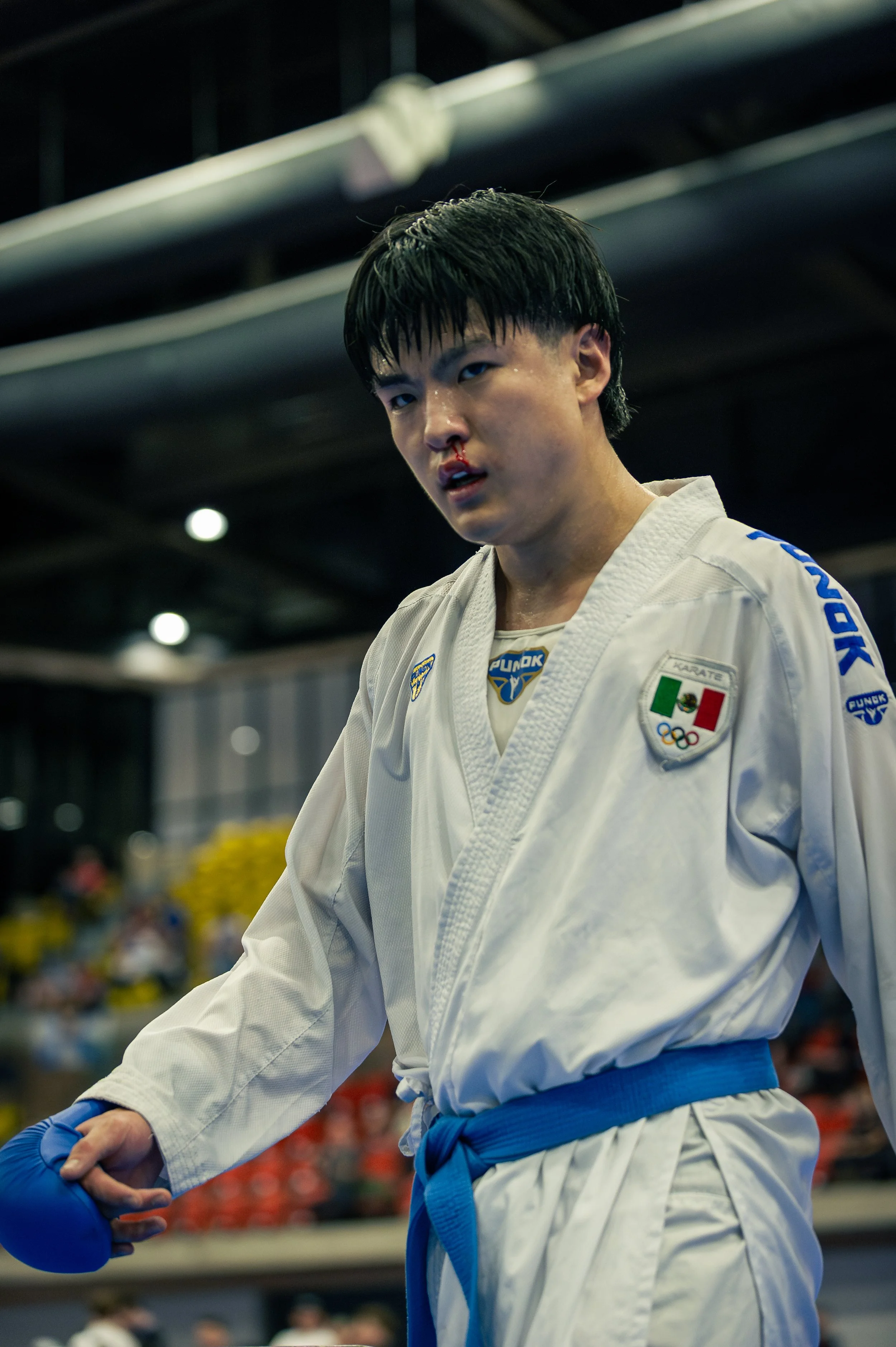 A young male martial artist wearing a white gi with blue belt and gloves, with a bloodied nose, representing mexico