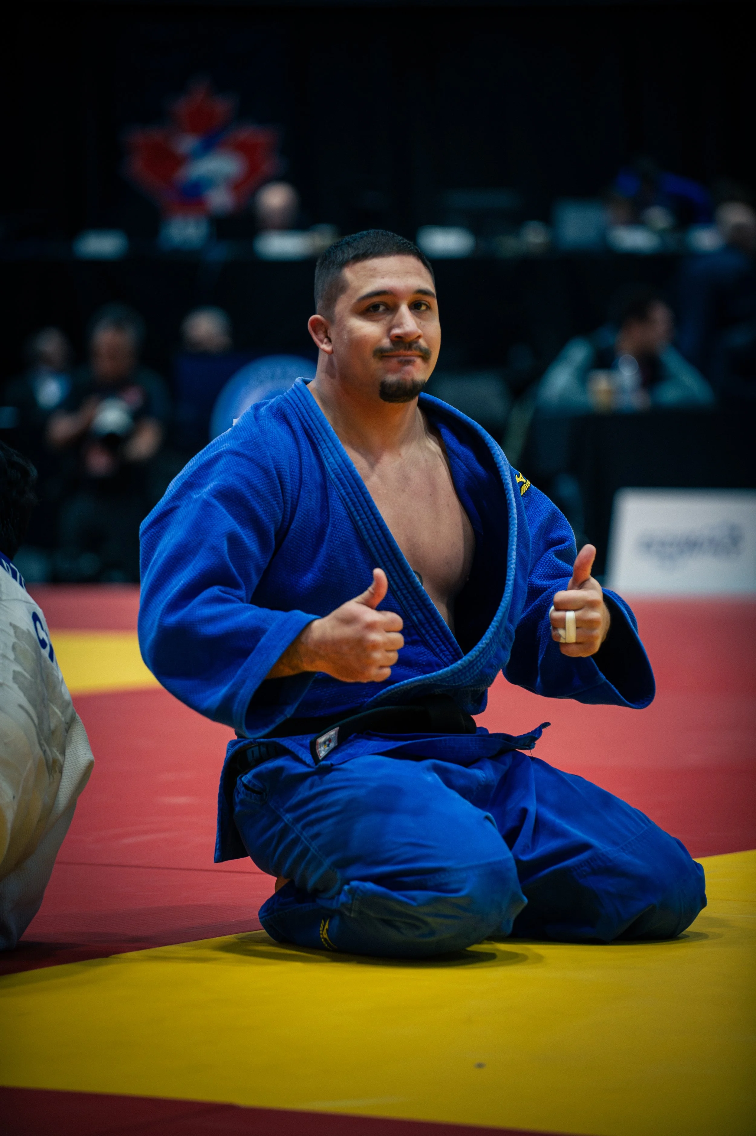 A male judo competitor kneeling on the yellow mat in a blue gi, giving a thumbs-up with both hands, with a confident expression. In the background, people are seated and a Canadian flag is visible.