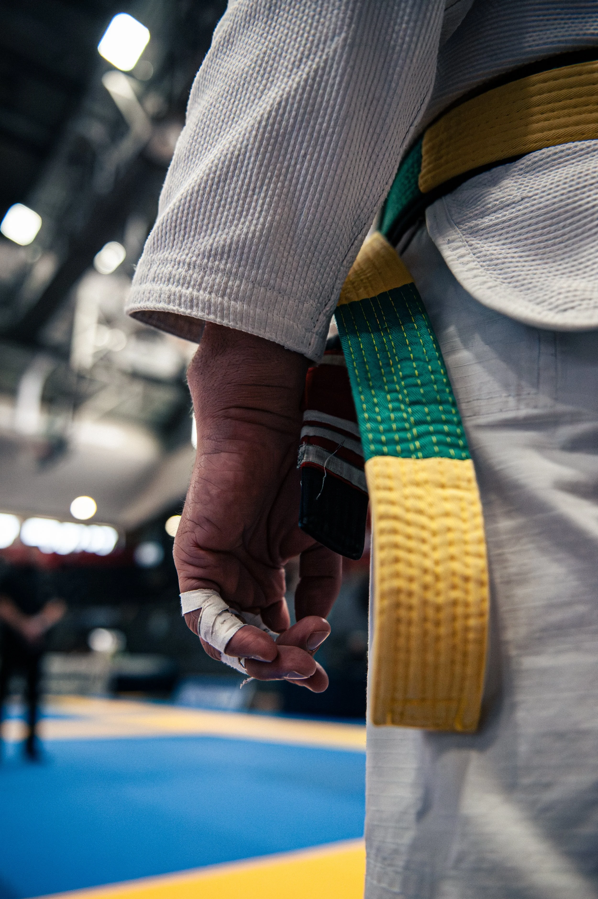 Close-up of a martial artist's hand with a bandage, in a white gi, holding a black and green belt, in a gym or dojo with a blue and yellow mat.