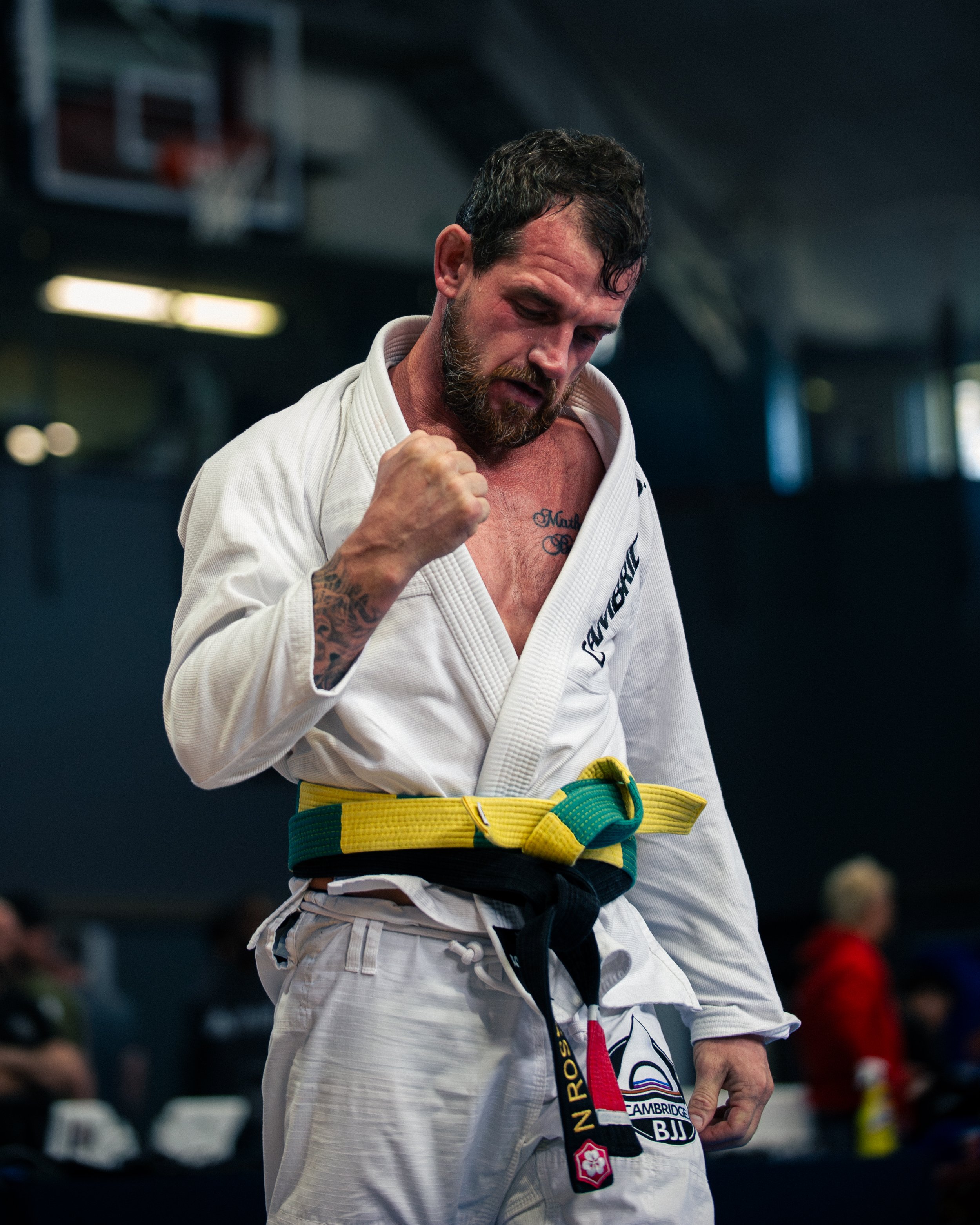 Man wearing a white Brazilian Jiu-Jitsu gi, adjusting a black  belt, in an indoor gym or dojo with blurred background.