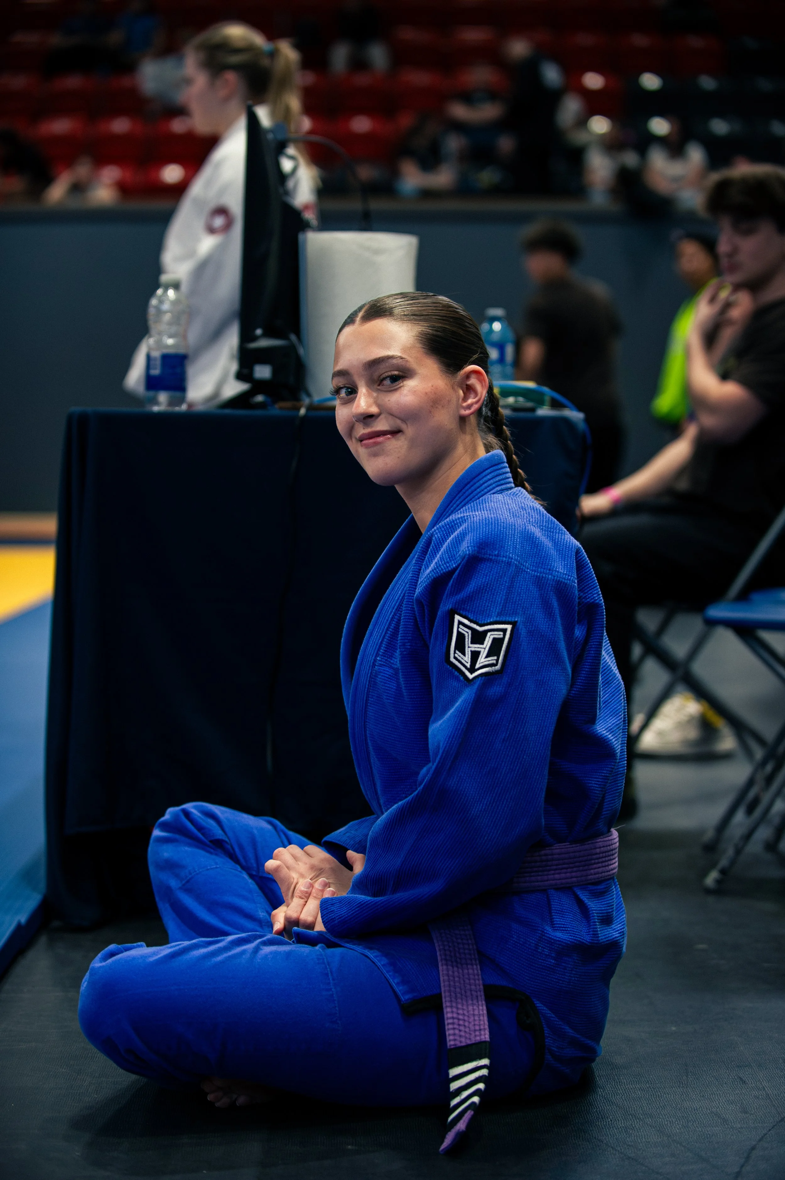 A young woman wearing a blue martial arts gi and purple belt sitting cross-legged on a black mat in a gym or competition setting. She is smiling at the camera with her hands resting on her knees, with a patch on her sleeve. In the background, other p