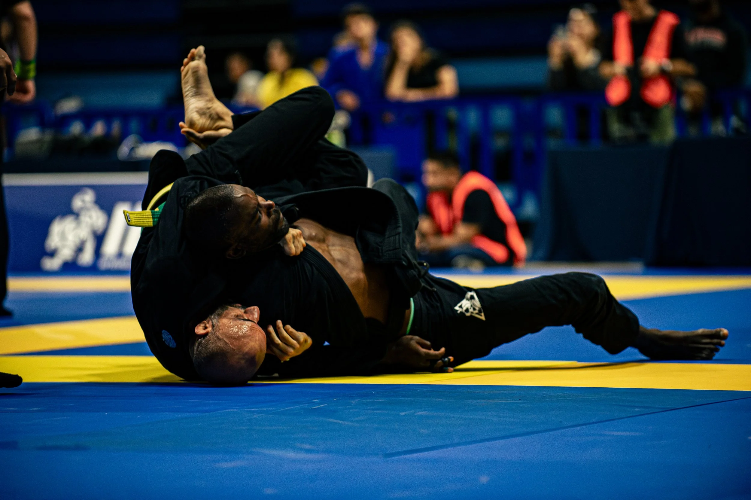 Two men in black martial arts uniforms practicing Brazilian Jiu-Jitsu techniques on a yellow and blue mat, with a blurred audience watching in the background.