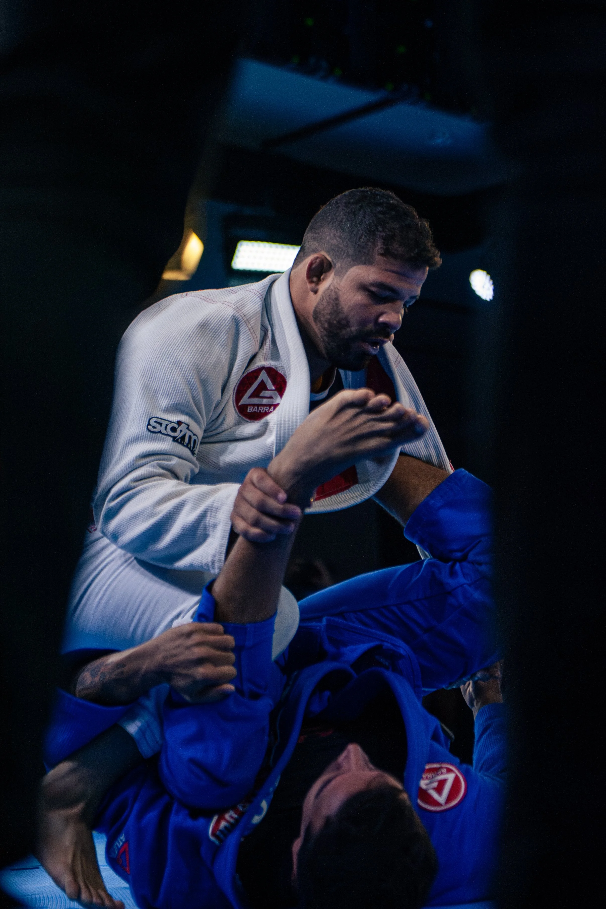 Two Brazilian Jiu-Jitsu competitors engaged in a grappling match, wearing rash guards with one person in a white gi with a red patch and the other in a blue gi. They are on a mat, surrounded by a dark environment with bright overhead lights.