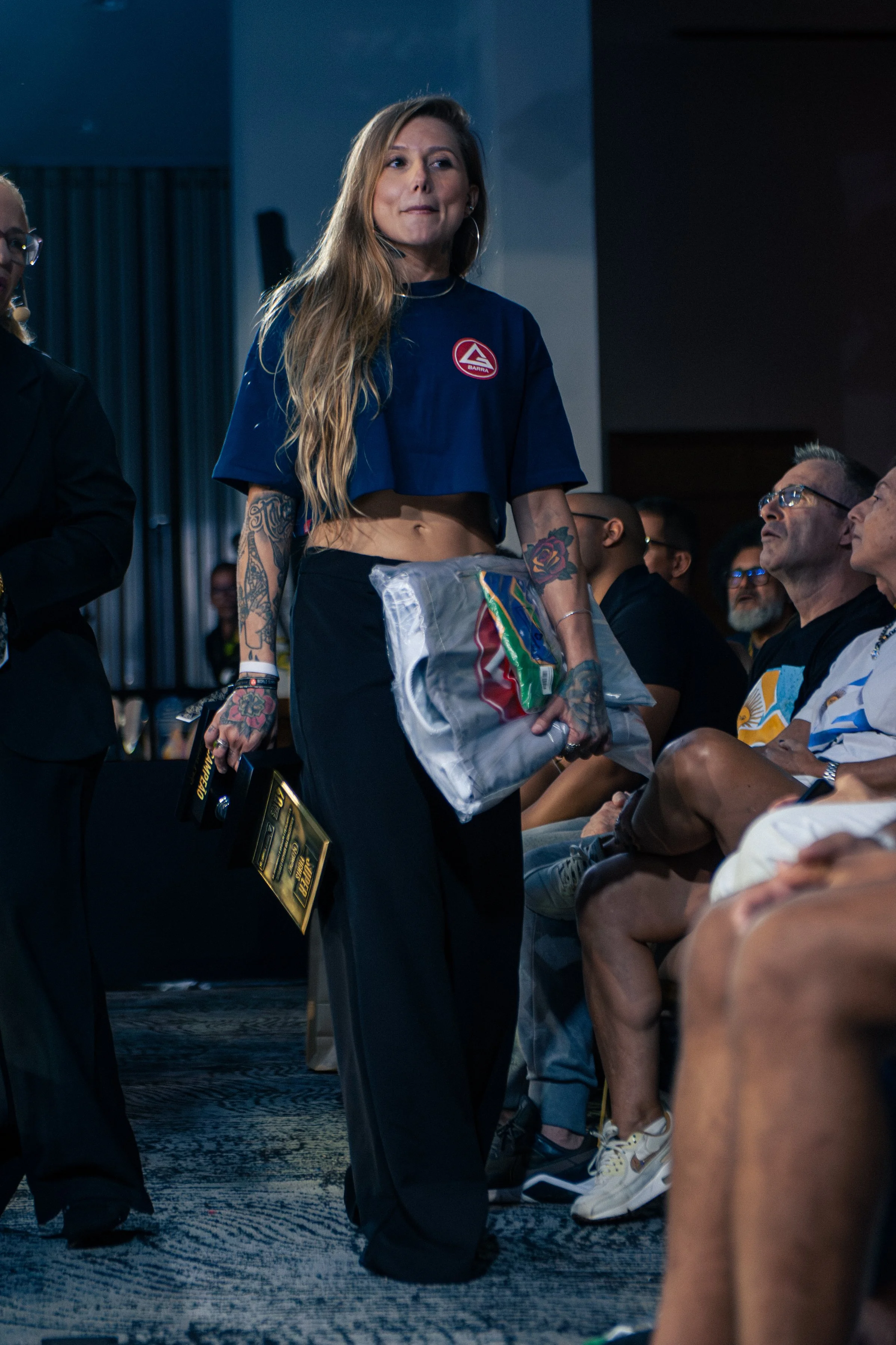 A woman with long wavy hair, tattoos on her arms, wearing a blue crop top with a logo, standing in a room with seated people.