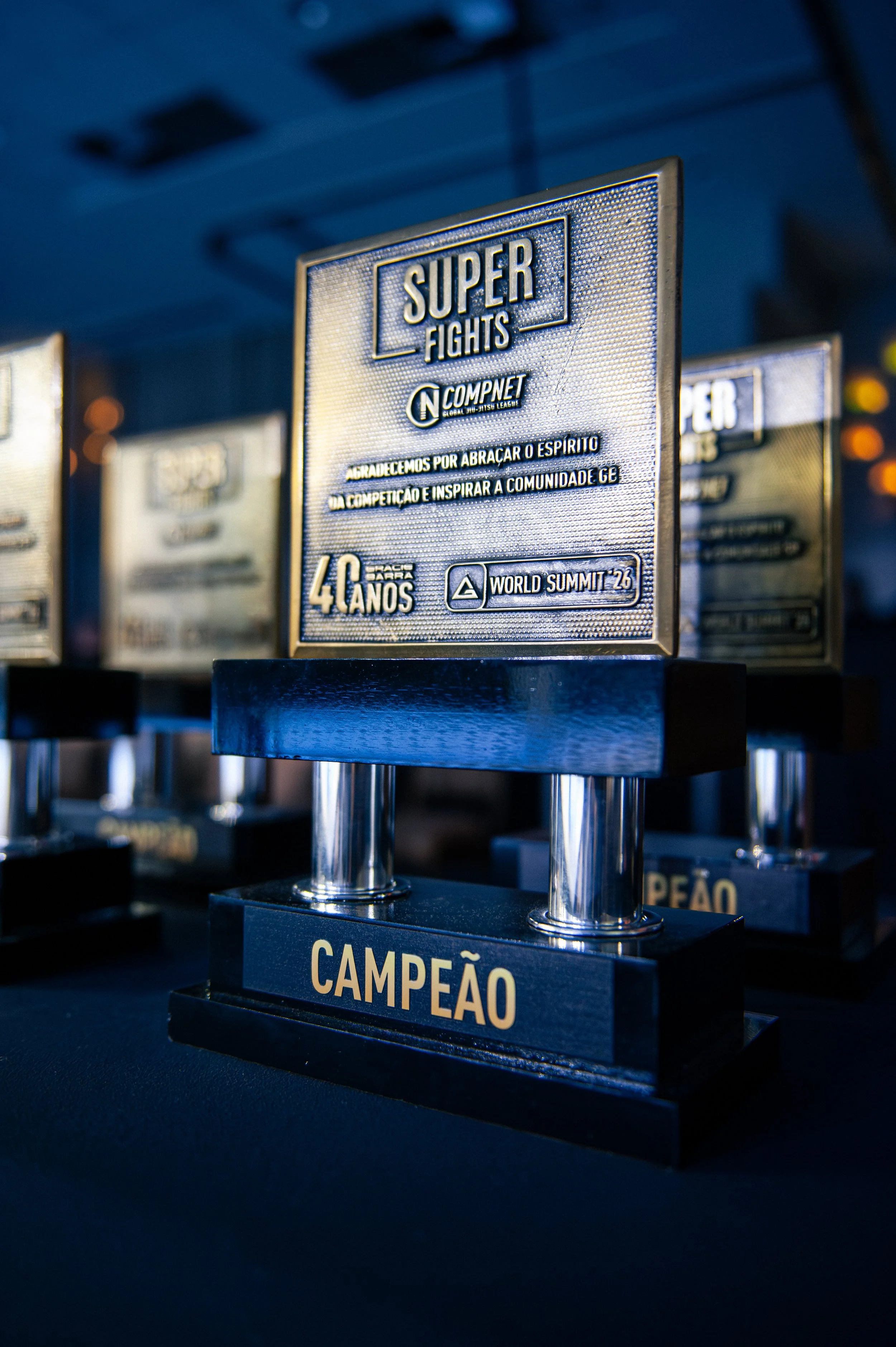 Trophies for a martial arts competition, with one prominently displaying the words 'Super Fights' and 'Campeão' on the base, and featuring a metallic plaque with Portuguese text expressing gratitude for inspiration and community impact.
